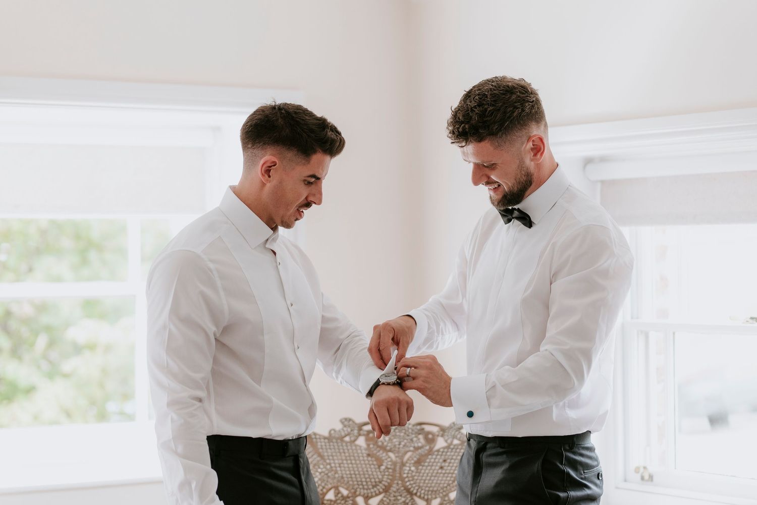 Two people in white dress shirts help each other with bow ties in a bright room with natural light.