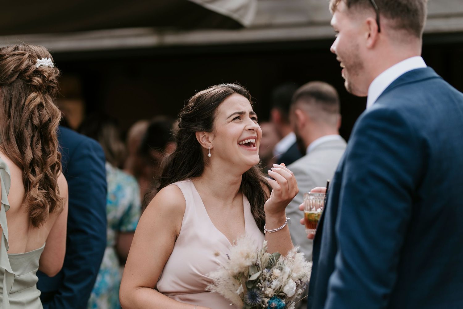 Wedding guests share a joyful moment while holding drinks at an outdoor reception gathering.