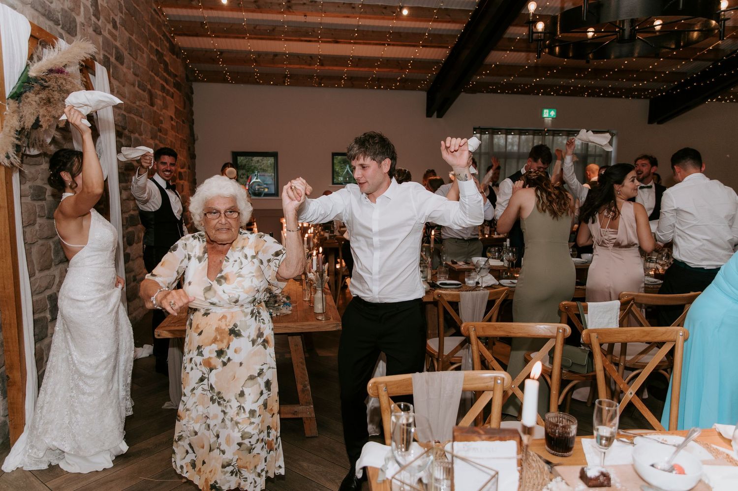 Wedding guests dancing and celebrating together in a rustic indoor venue with exposed brick walls and wooden chairs.