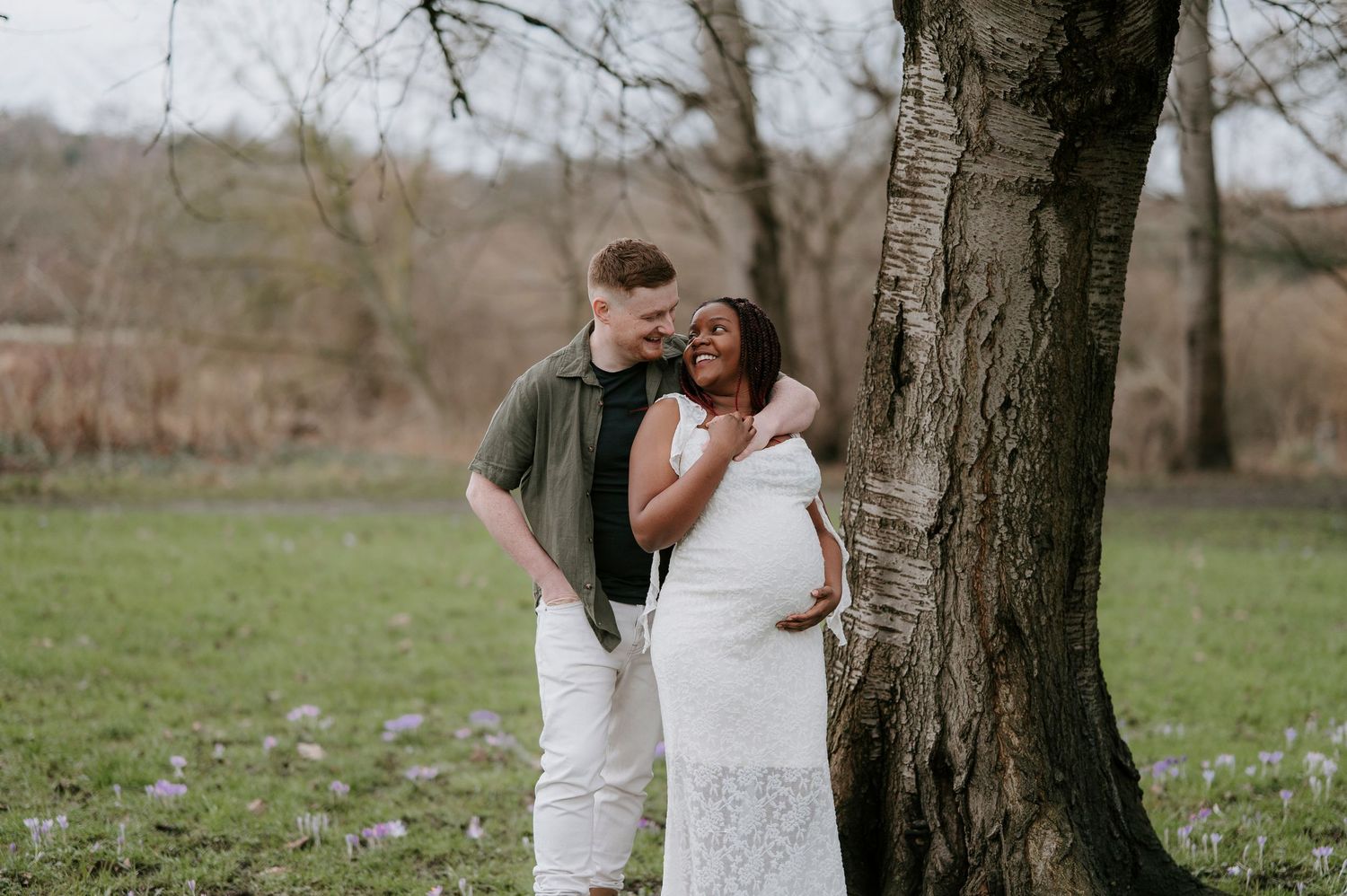 A couple shares a romantic moment under a tree in a spring meadow with small purple wildflowers blooming.