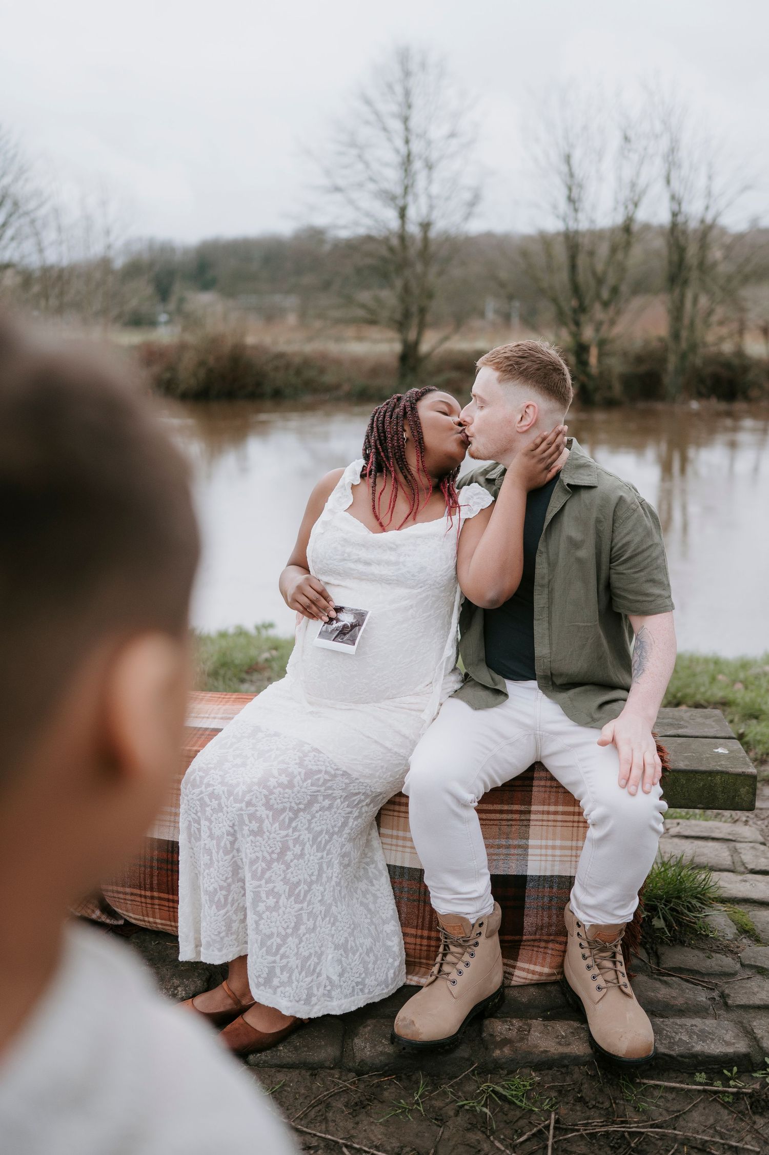 Romantic couple sharing an intimate moment while sitting on a wooden bench by a serene lake on a cloudy day.