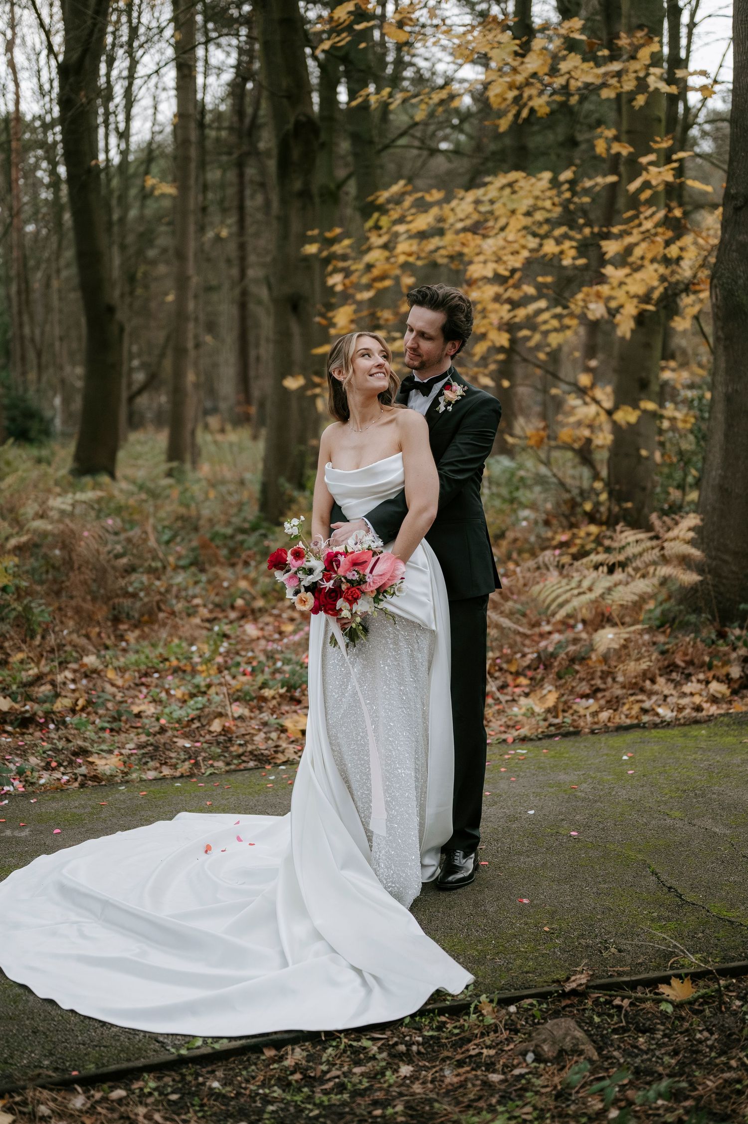 Elegant bride in flowing white gown laughs joyfully with groom among autumn trees.