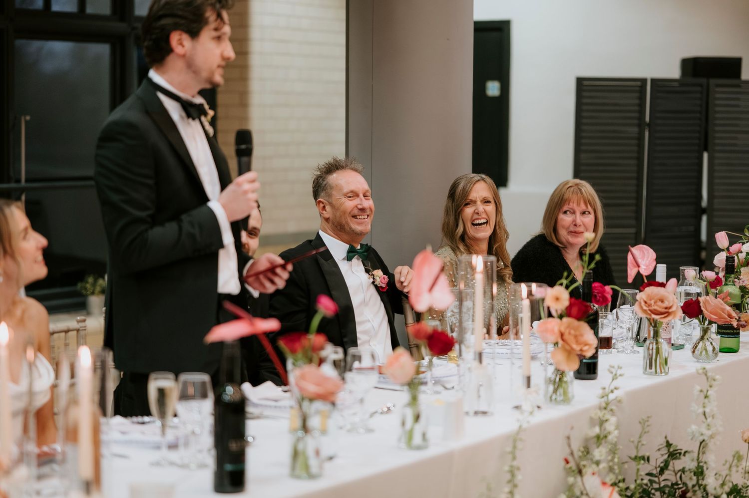 Wedding guests at a reception table laugh and smile during a toast while surrounded by floral centerpieces and glassware.