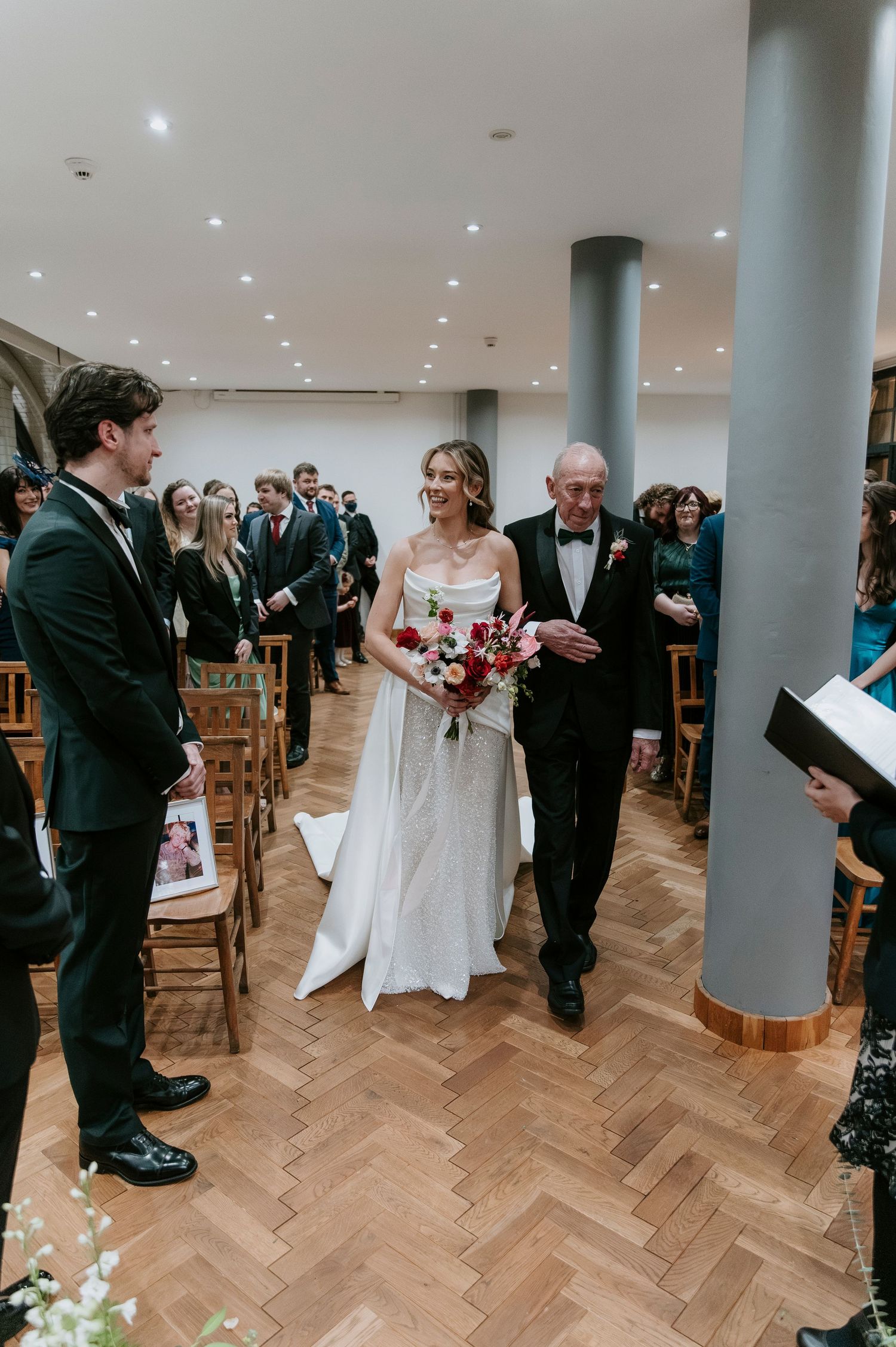 Wedding ceremony in a modern venue with herringbone wood floors and white pillars showing guests seated in wooden chairs.