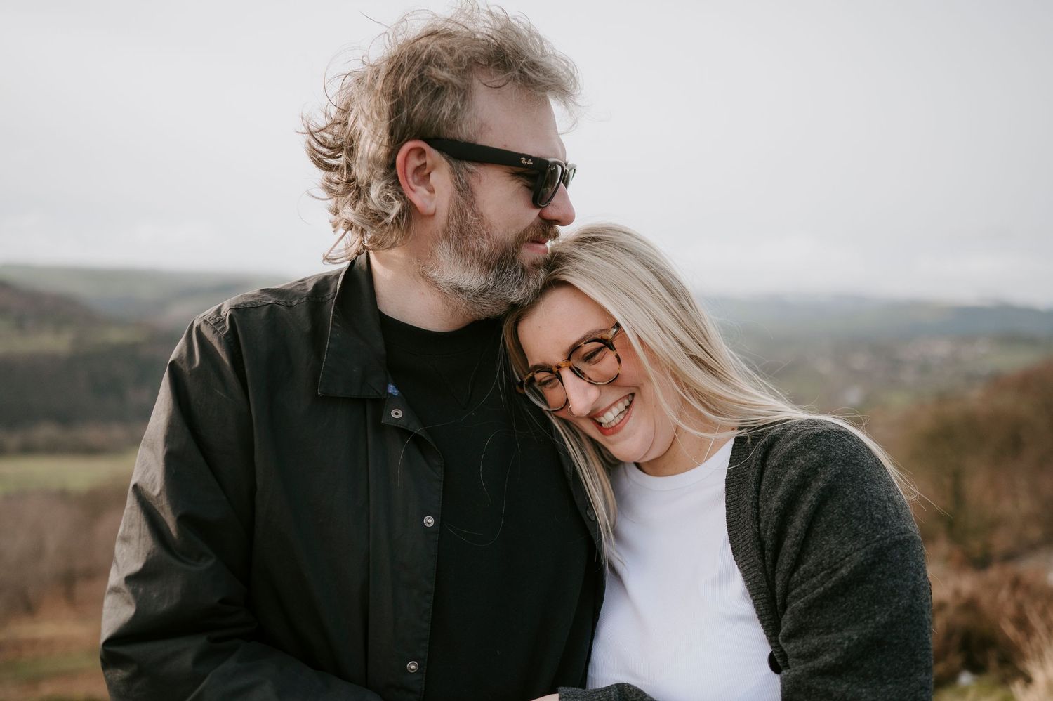 A couple in casual attire embraces while standing in a scenic outdoor setting with rolling hills in the background.