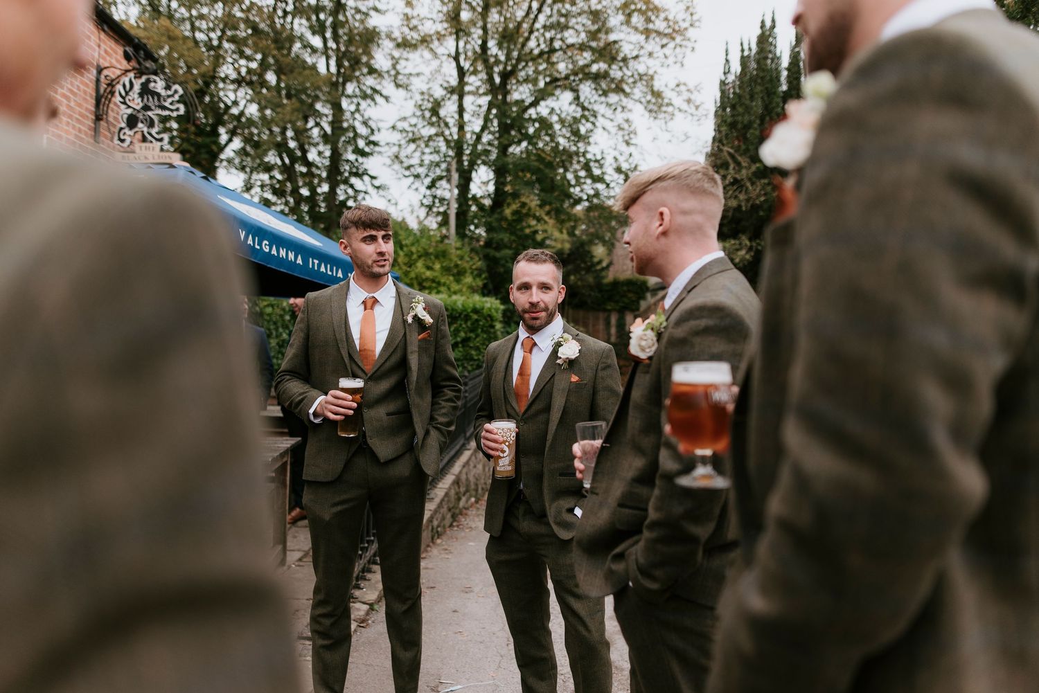Wedding guests in formal attire gather outdoors near a brick building for a social event.