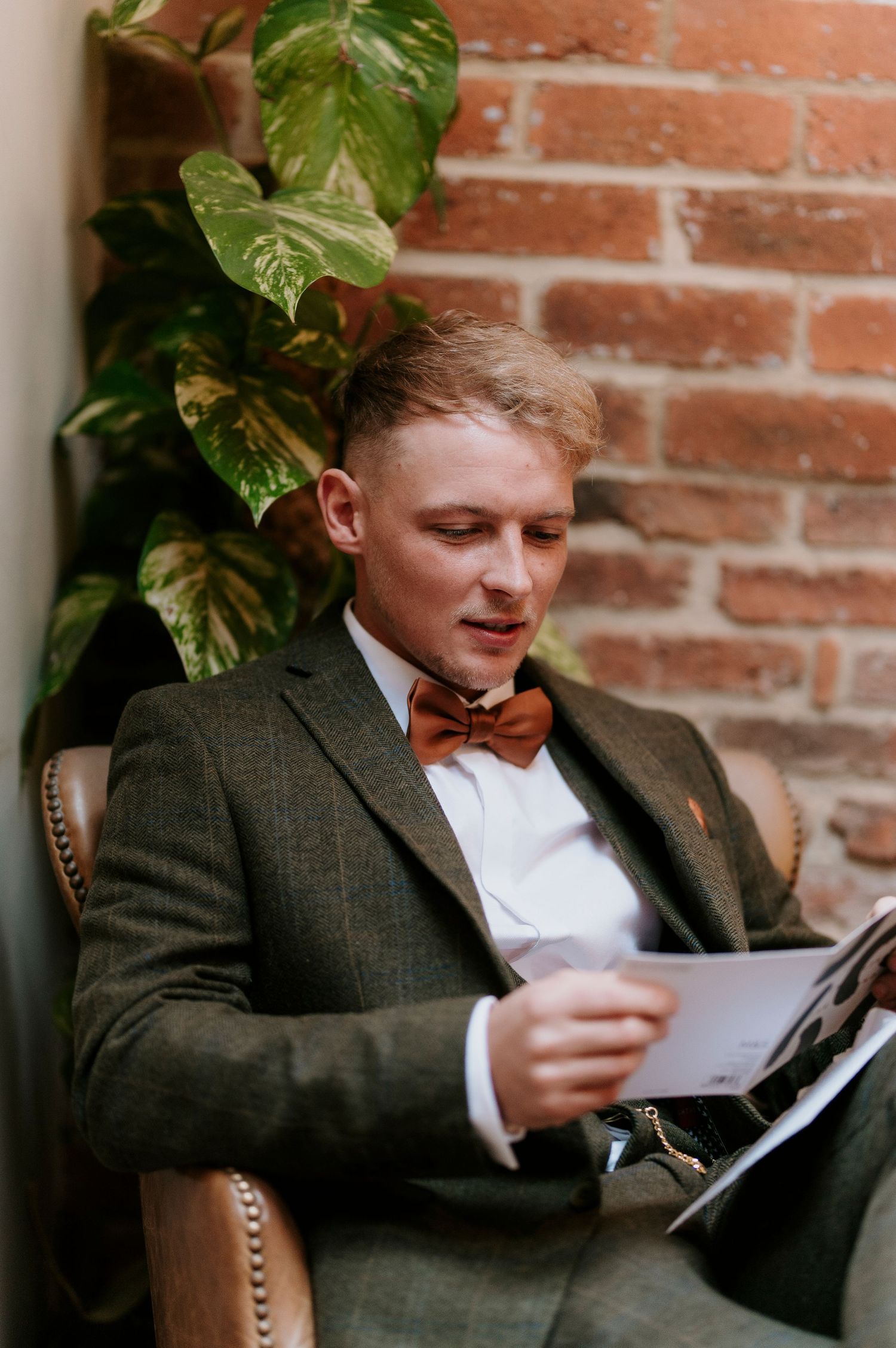 Person in tweed jacket reads papers while sitting against brick wall with green foliage.