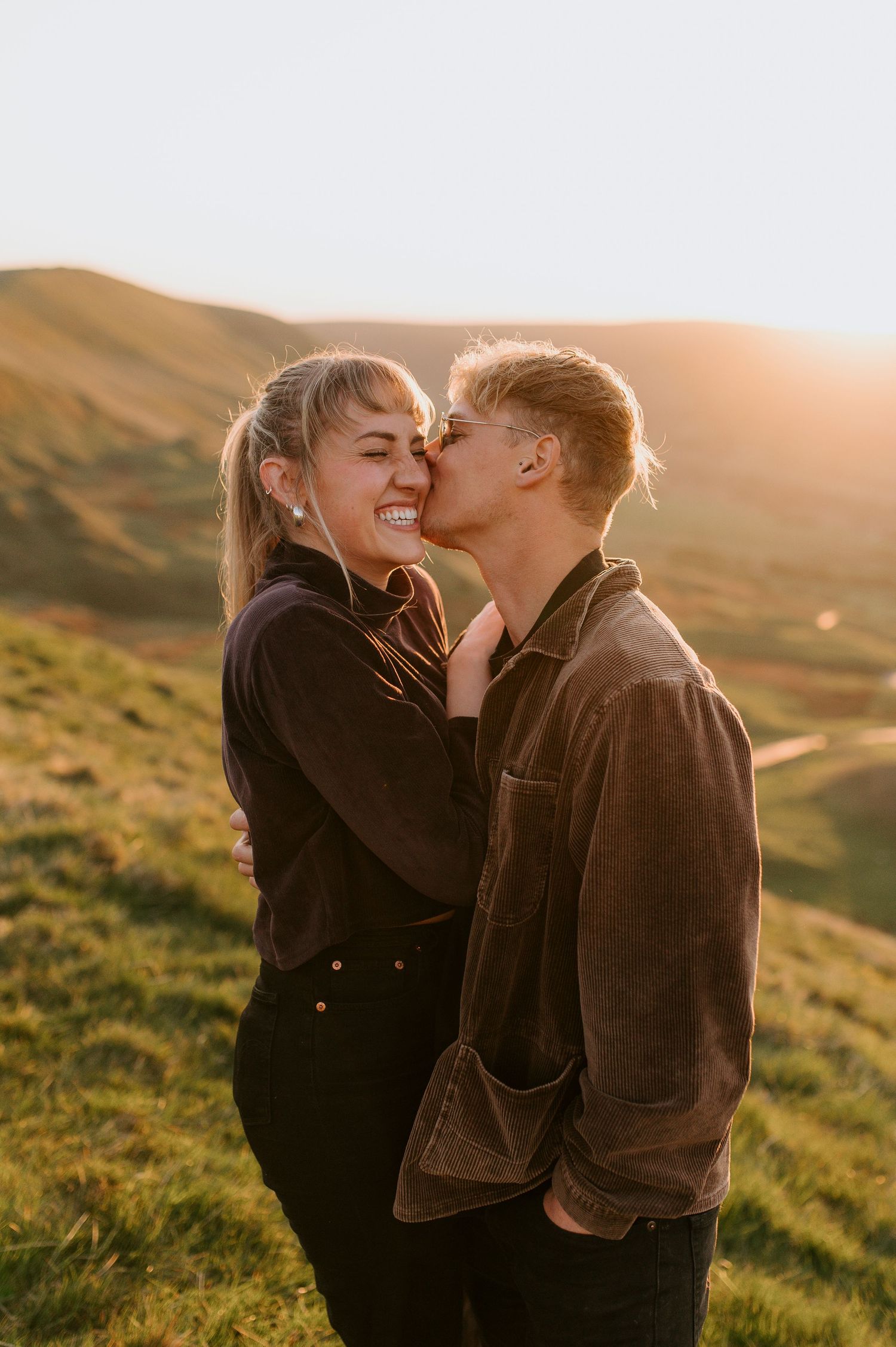 A romantic couple shares an intimate moment at sunset on a grassy hillside in warm golden light.