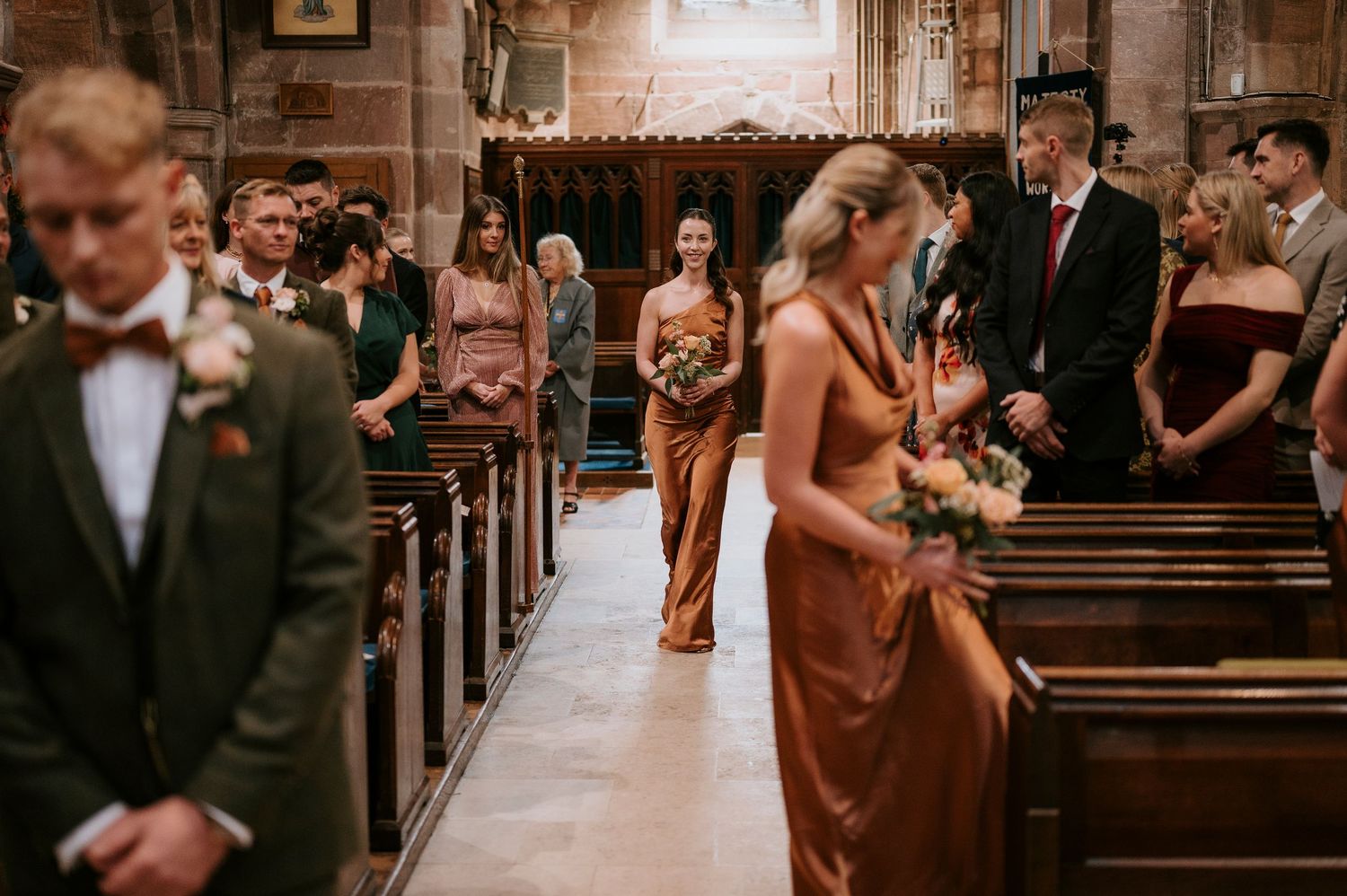 Wedding party members in rust-colored dresses walk down the aisle of a traditional stone church during a ceremony.