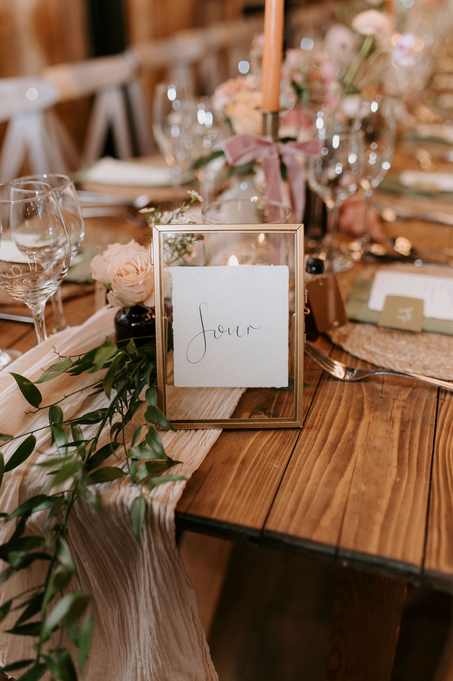 Elegant wedding table setting with gold framed table number, blush flowers, and greenery on wooden farmhouse table.