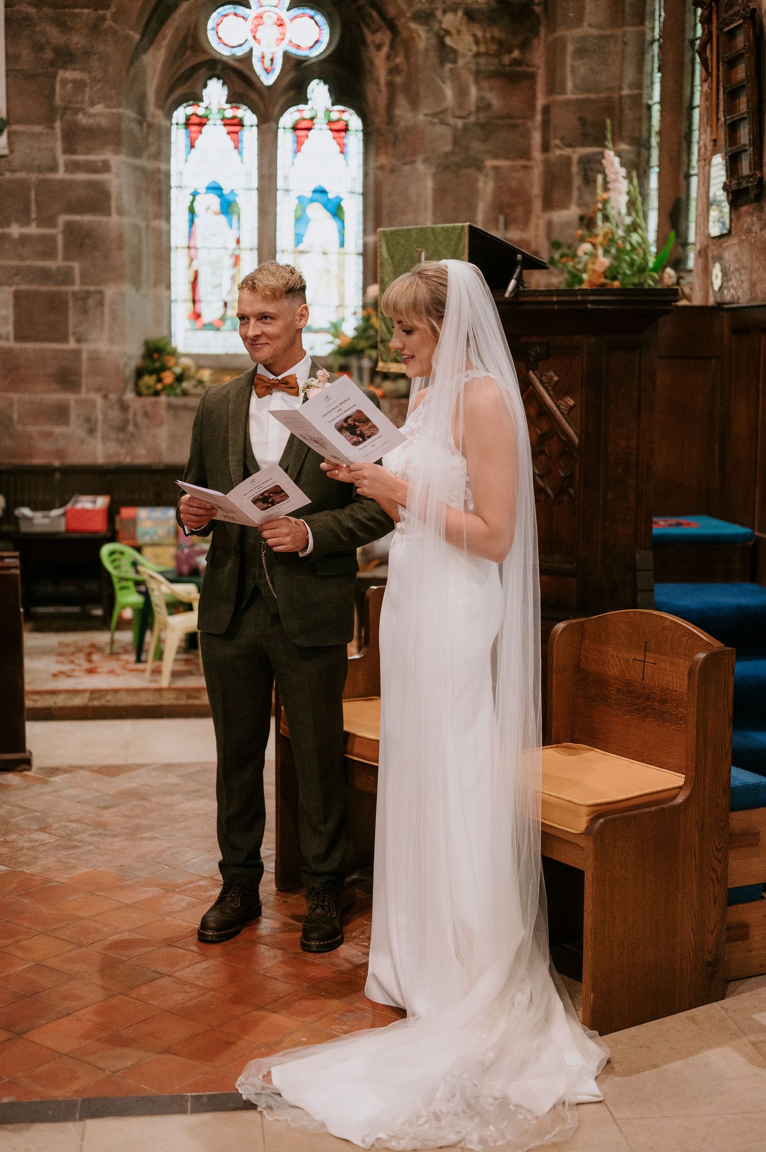 A bride and groom stand together reading from ceremony programs inside a stone church with stained glass windows.
