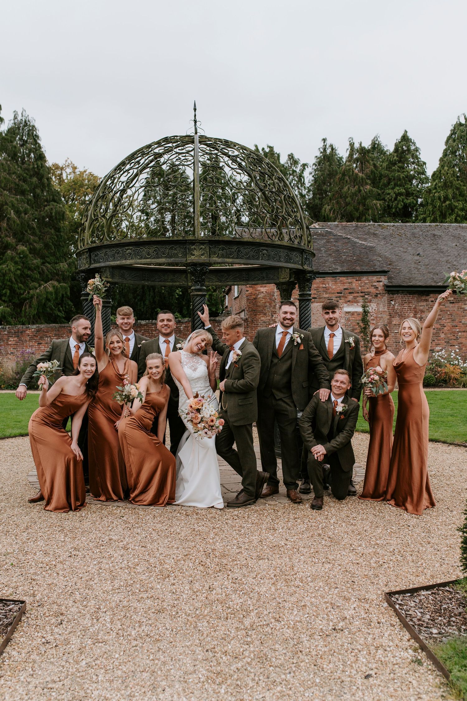 Wedding party poses together on gravel path in front of rustic brick wall and garden archway.