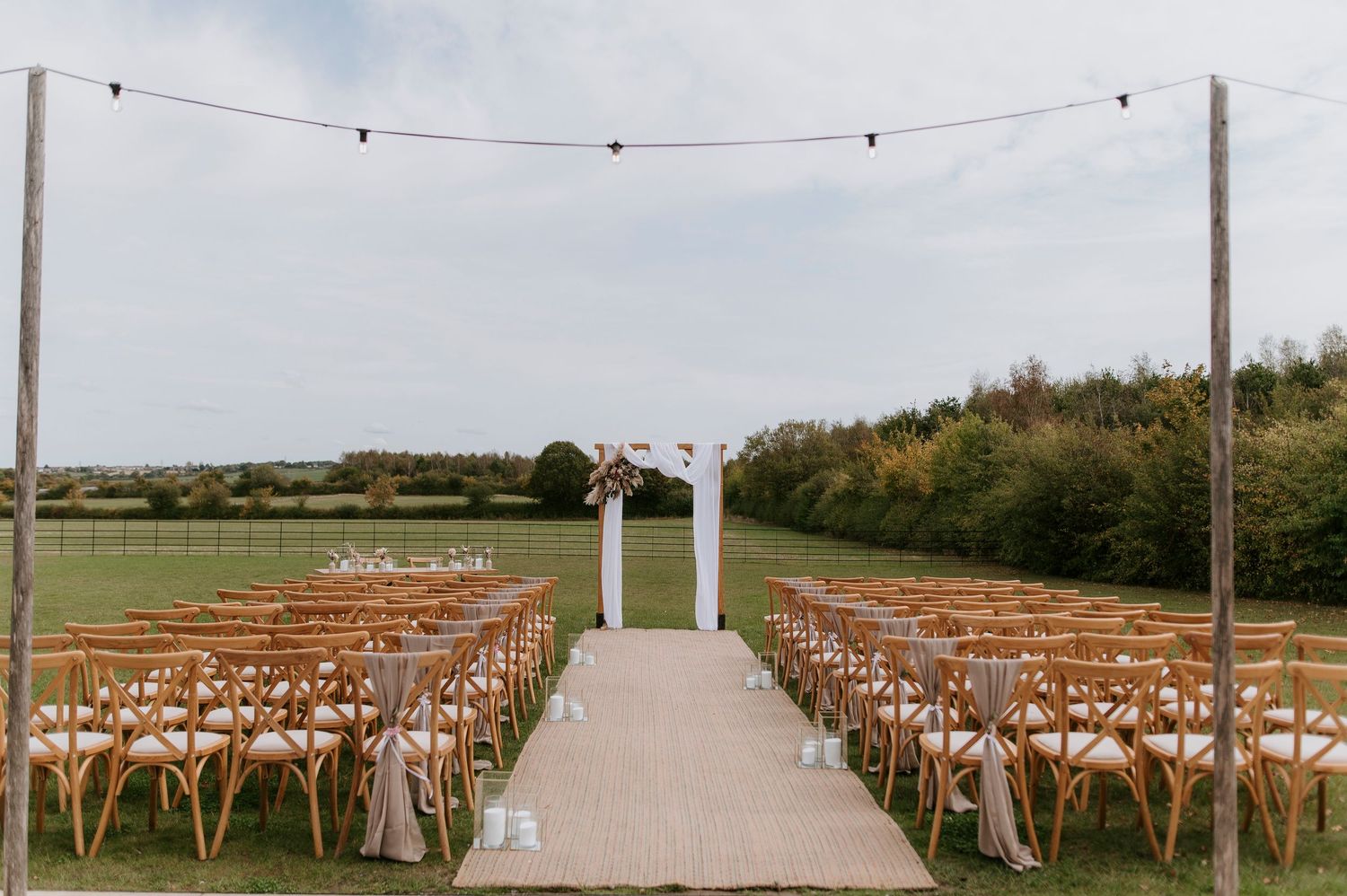 Outdoor wedding ceremony setup with wooden chairs and string lights along a path in a rustic countryside setting.