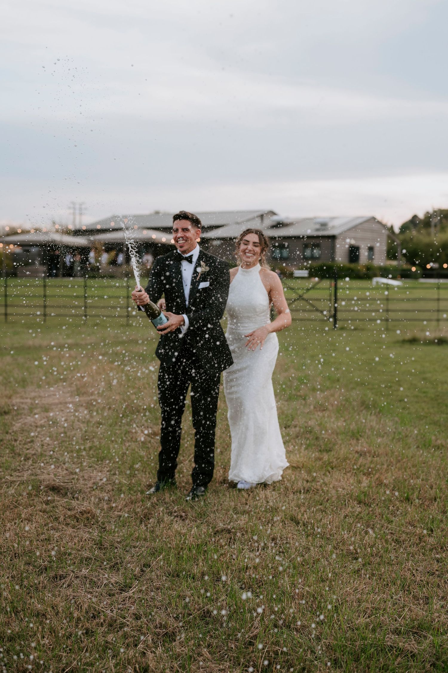 A couple pops a champagne bottle in celebration while standing in a grassy field on an overcast day.