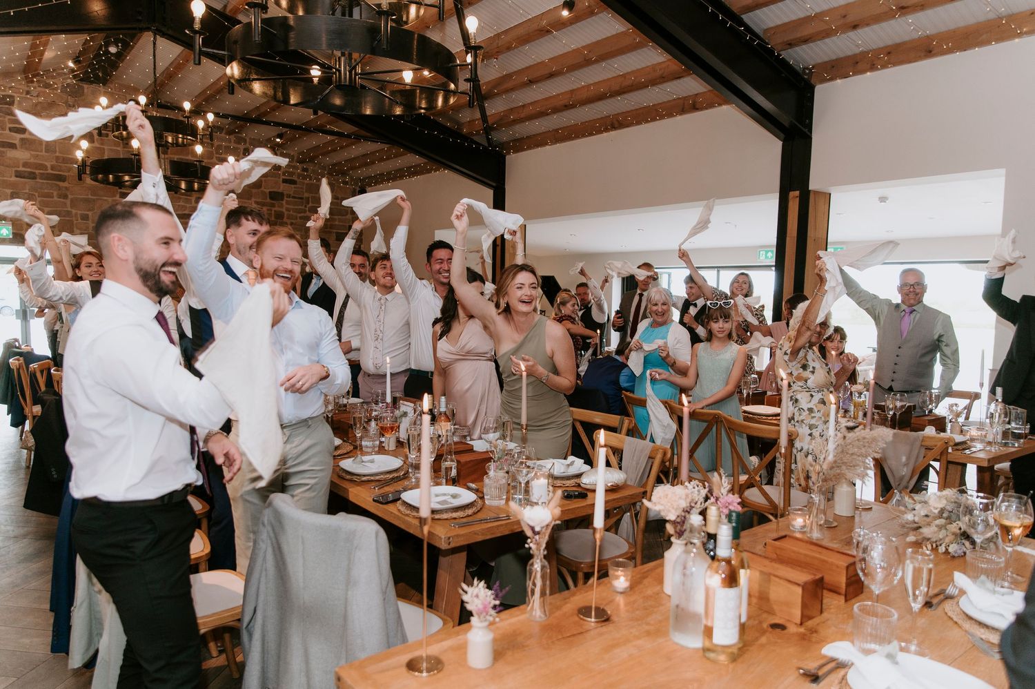 Wedding guests enjoy a lively reception dinner at long wooden tables in a rustic venue with exposed beam ceilings.