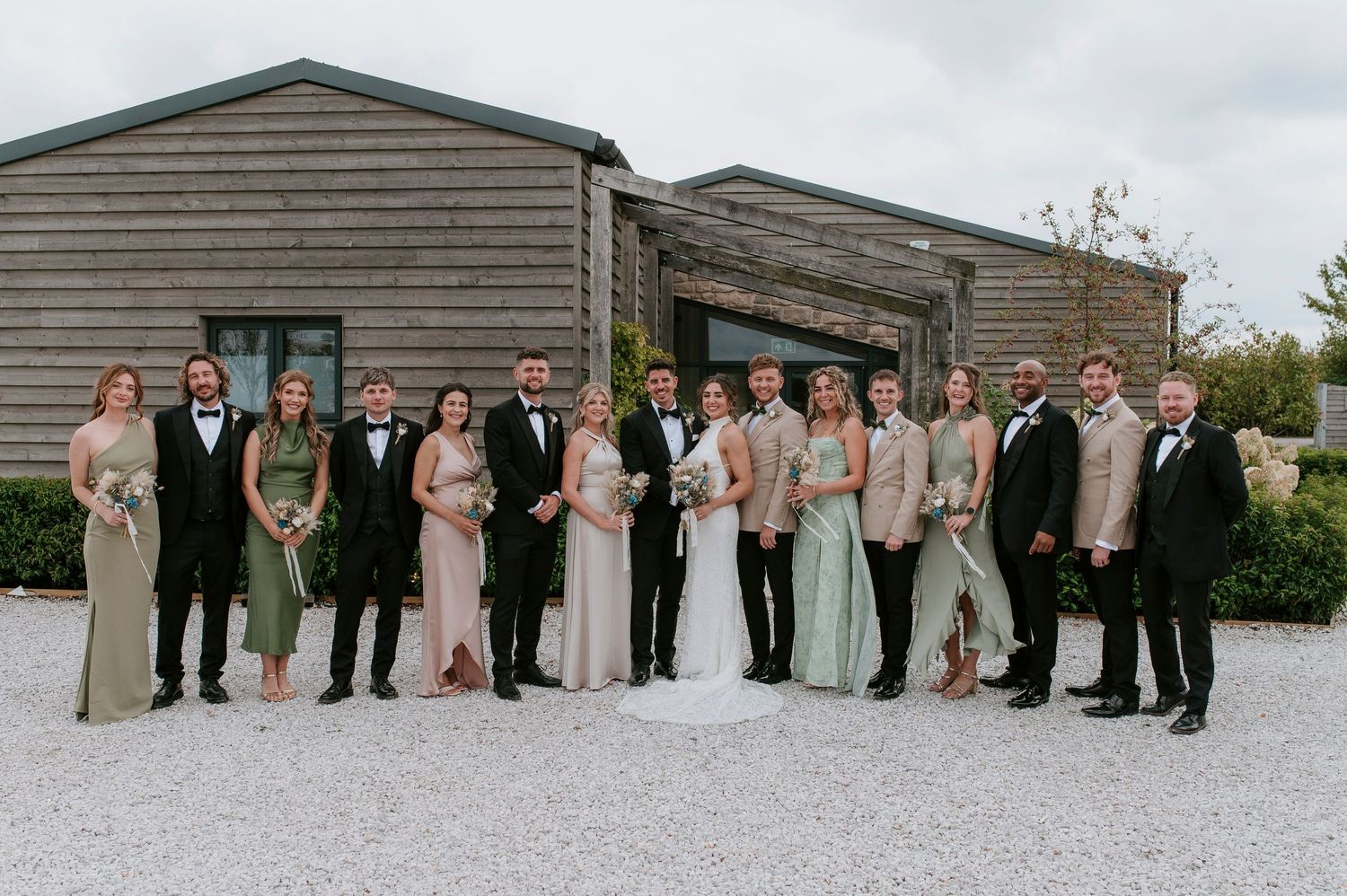 Wedding party members stand in a line outside a rustic wooden building during an outdoor celebration.