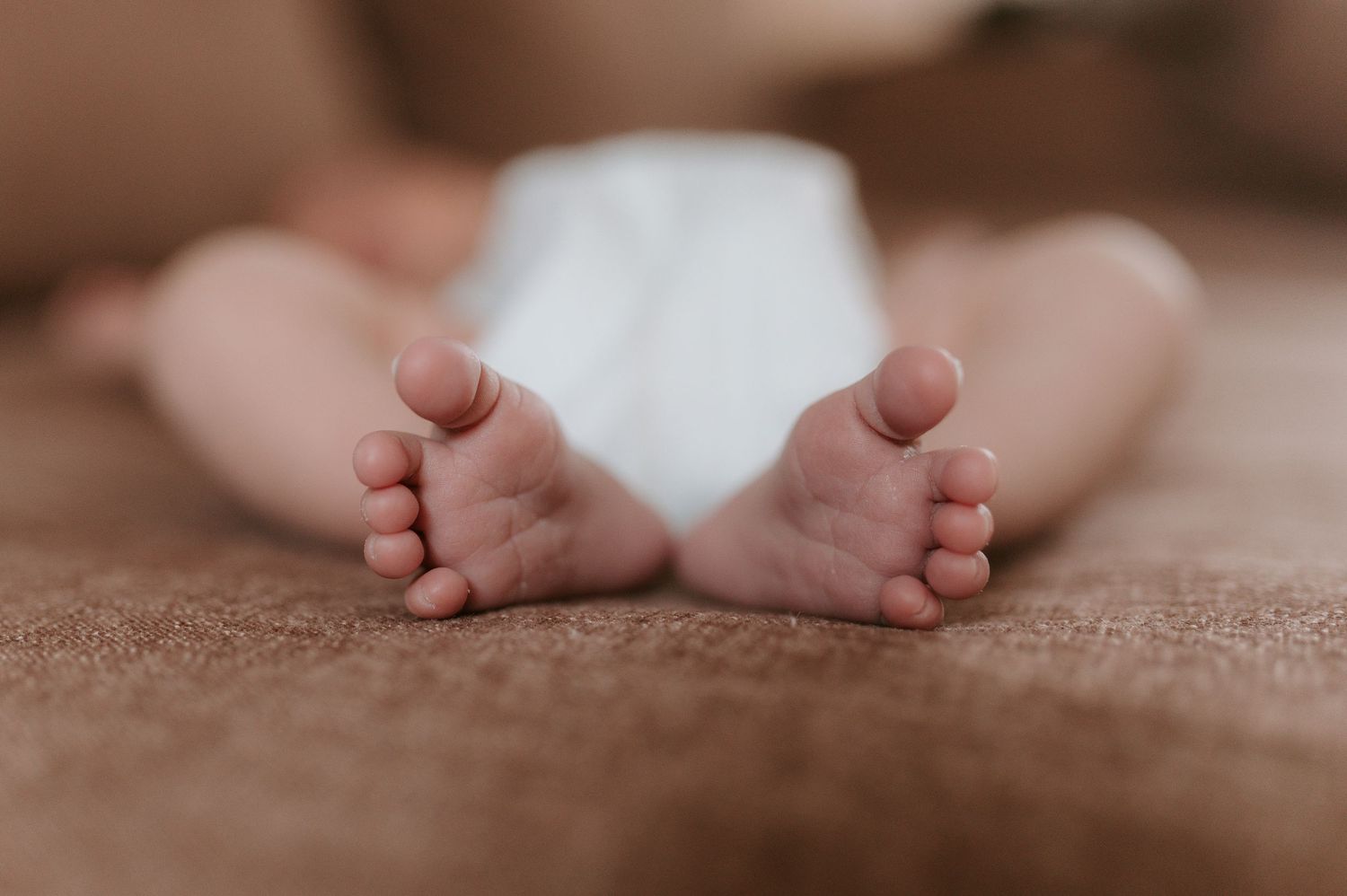 Close-up of tiny baby feet resting on soft brown carpet showing delicate toes and soles.
