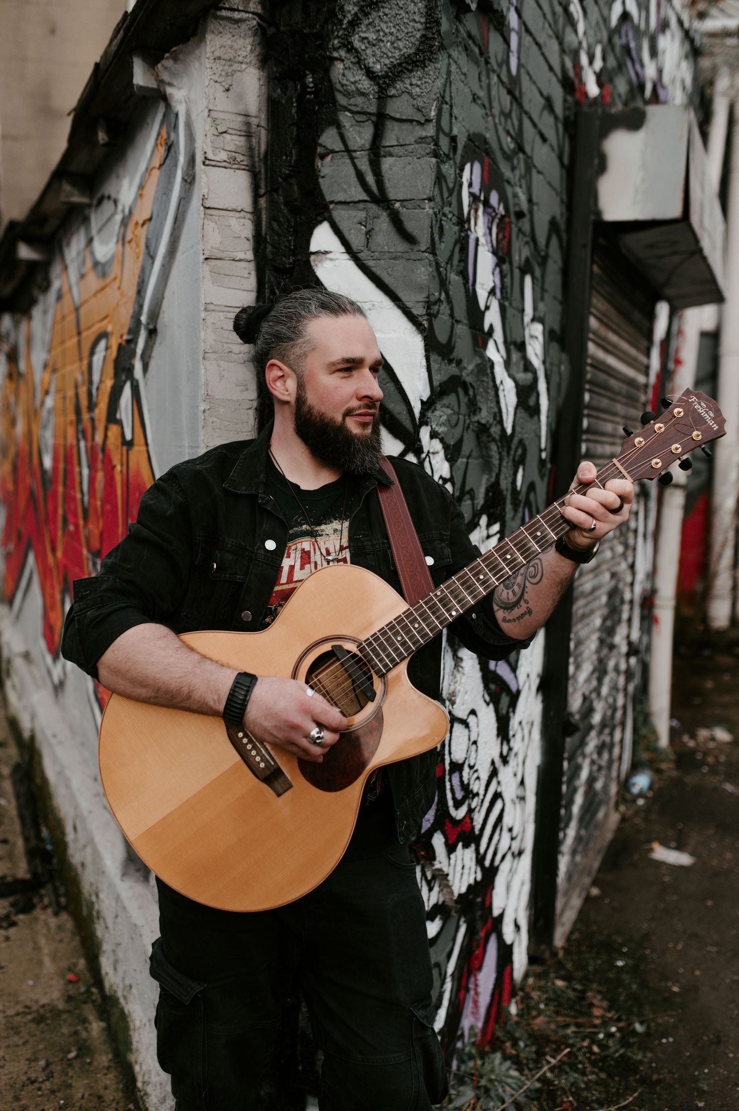 Musician performing with acoustic guitar against graffiti covered wall in urban setting.