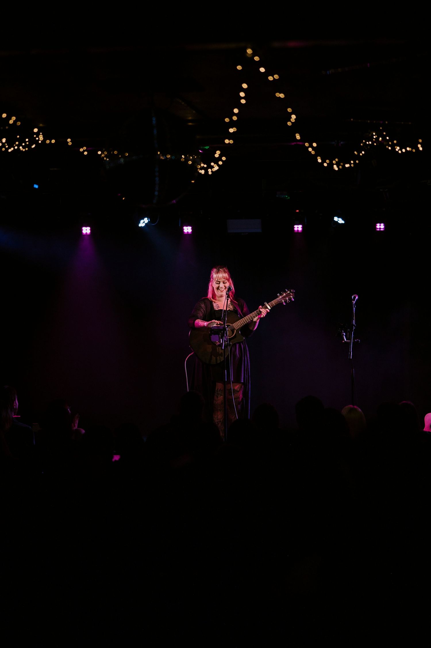 A musician performs on stage with an acoustic guitar under string lights and purple stage lighting.
