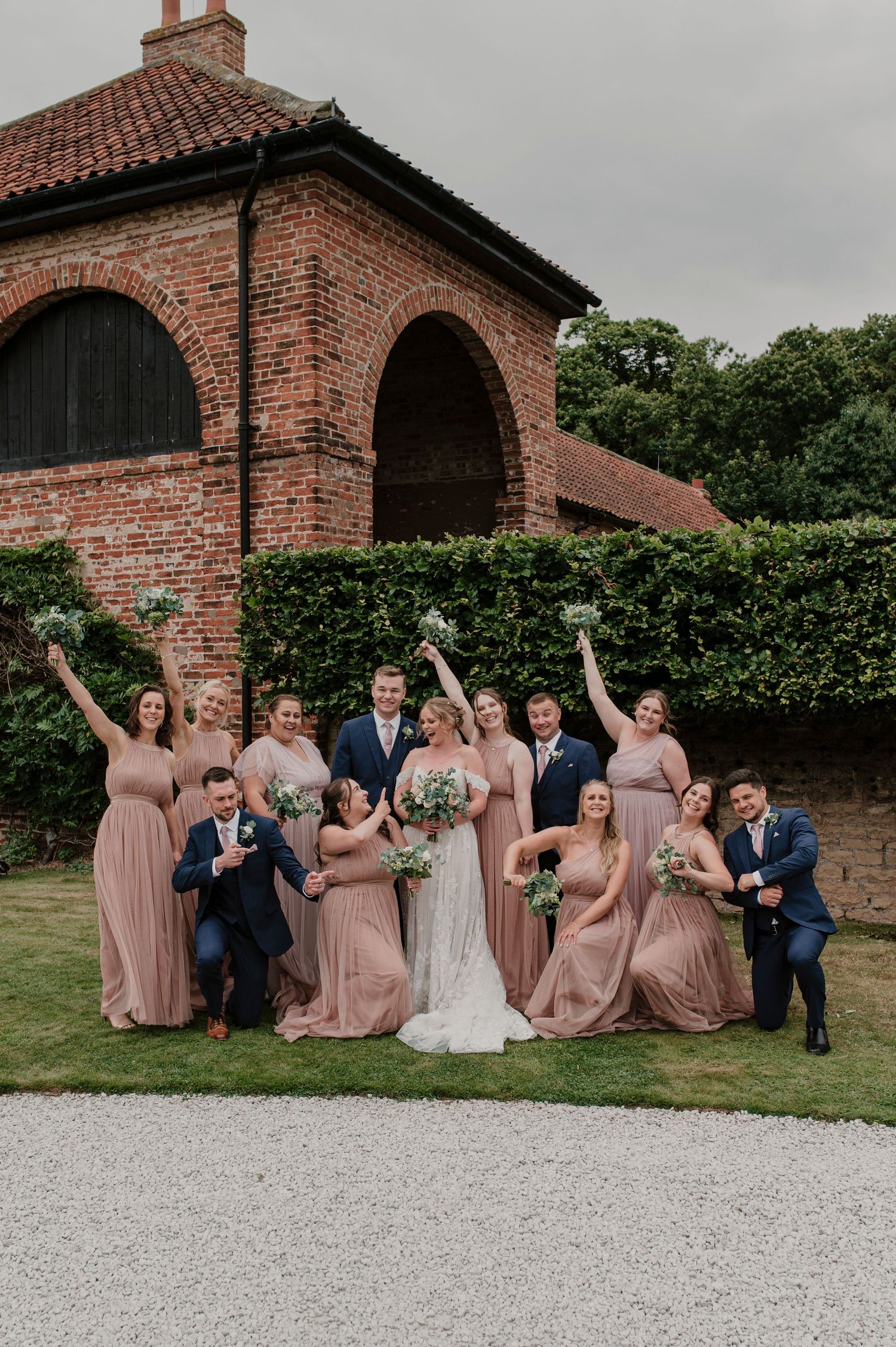 Wedding party in dusty rose dresses and navy suits pose playfully against brick wall and hedges.