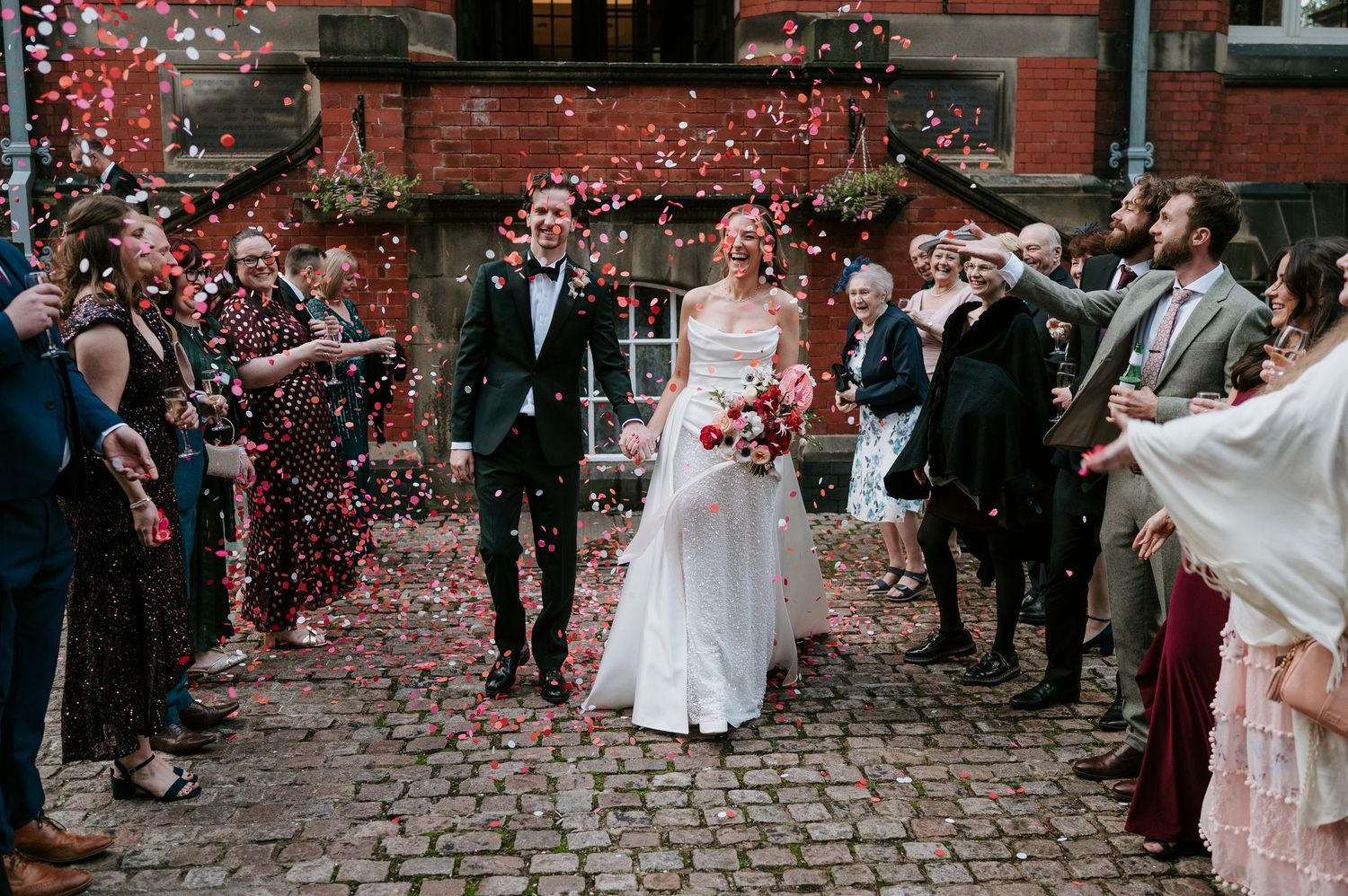 Guests shower newlyweds with rose petals as they walk down a cobblestone path outside a brick building.
