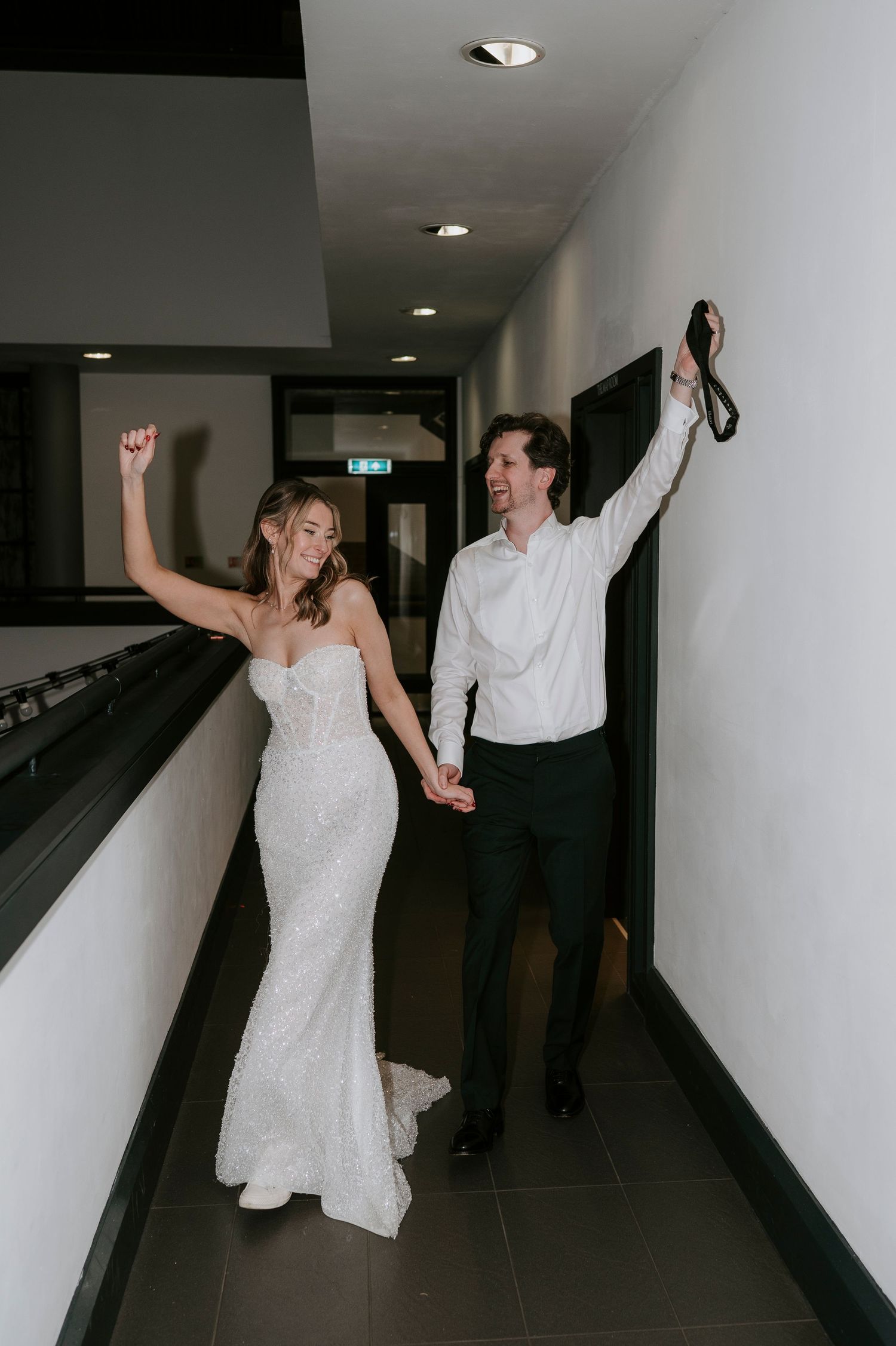 A couple in wedding attire playfully dances and poses together in a dimly lit hallway corridor.