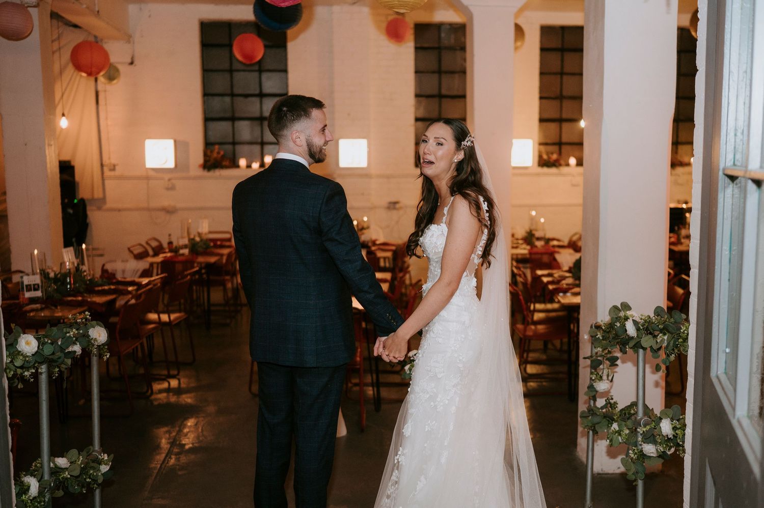 Bride in lace gown and groom in navy suit hold hands and smile in elegant industrial wedding reception hall.
