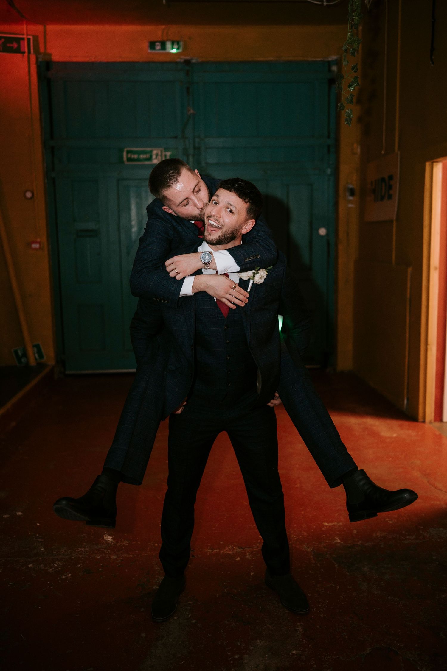 Two groomsmen in navy suits share a fun piggyback ride moment at a wedding celebration.