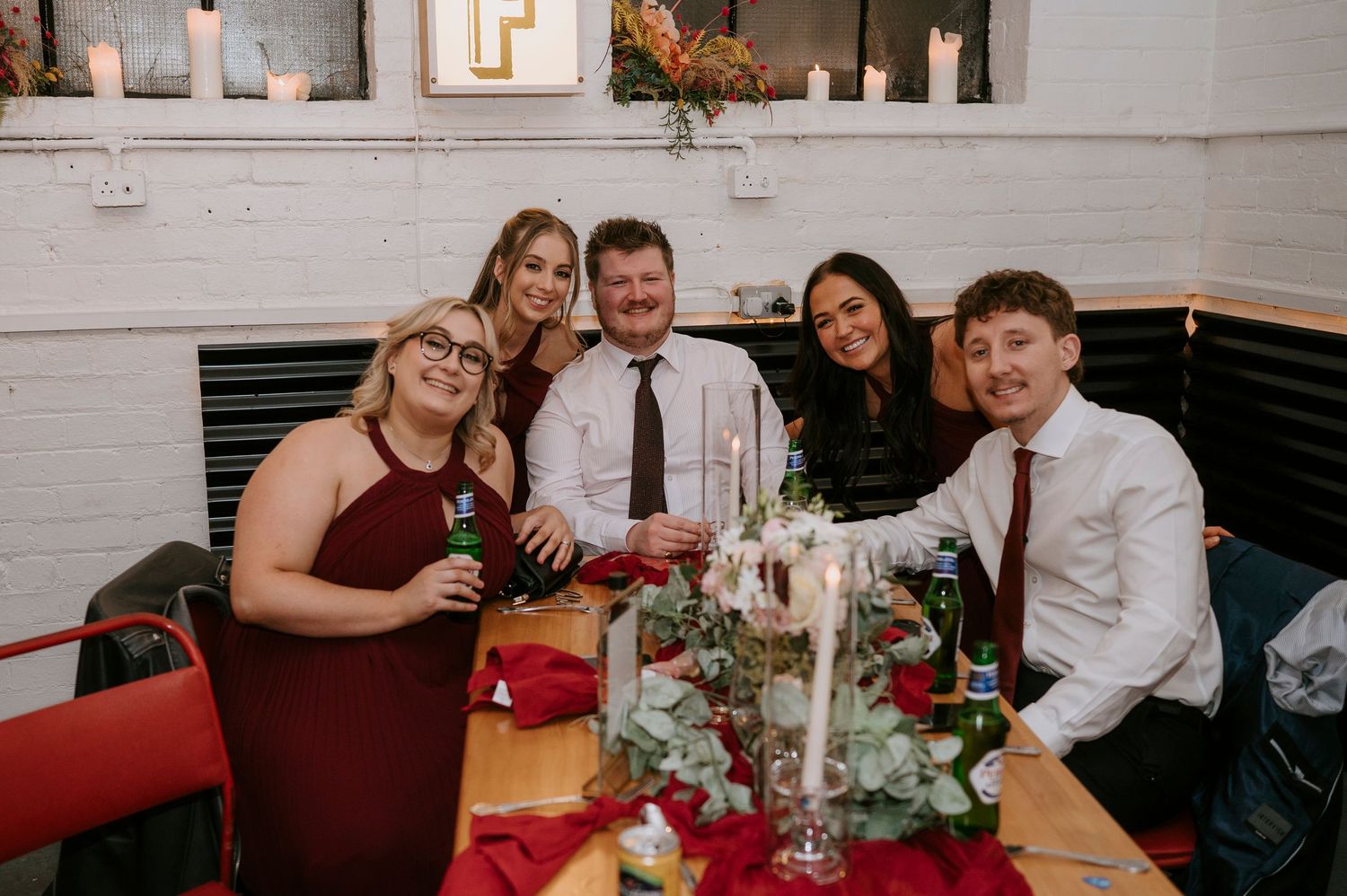 Group of five wedding guests smile at decorated reception table with red roses and champagne glasses.