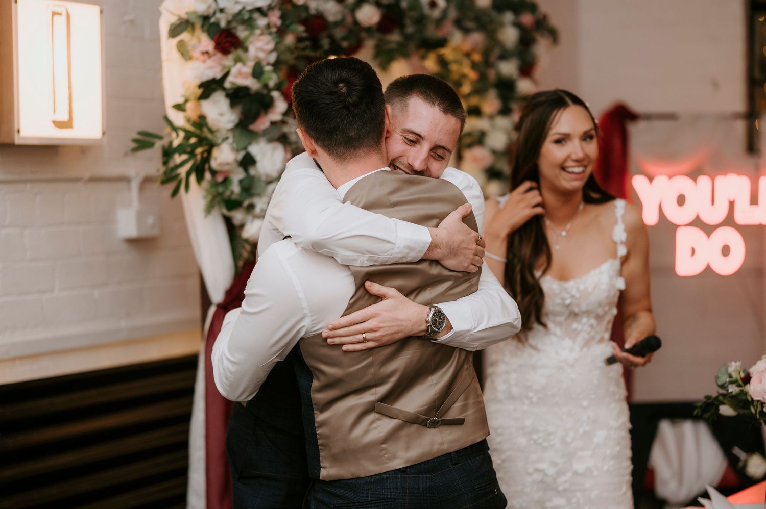Groom hugging guest at wedding reception while bride smiles near You'll Do neon sign.