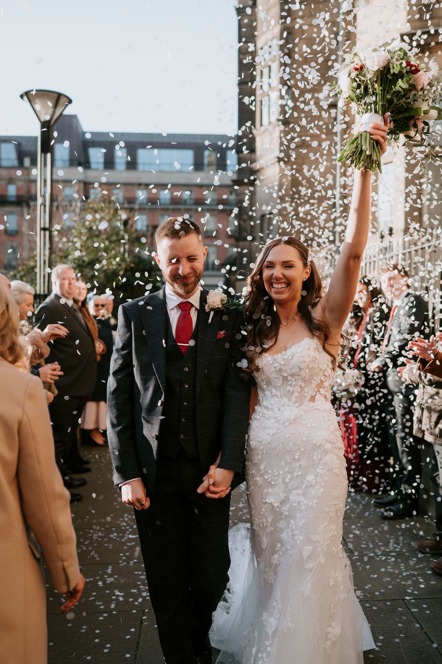 Bride and groom walk through confetti shower, bride raising bouquet, groom in dark suit with red tie, celebrating wedding exit.