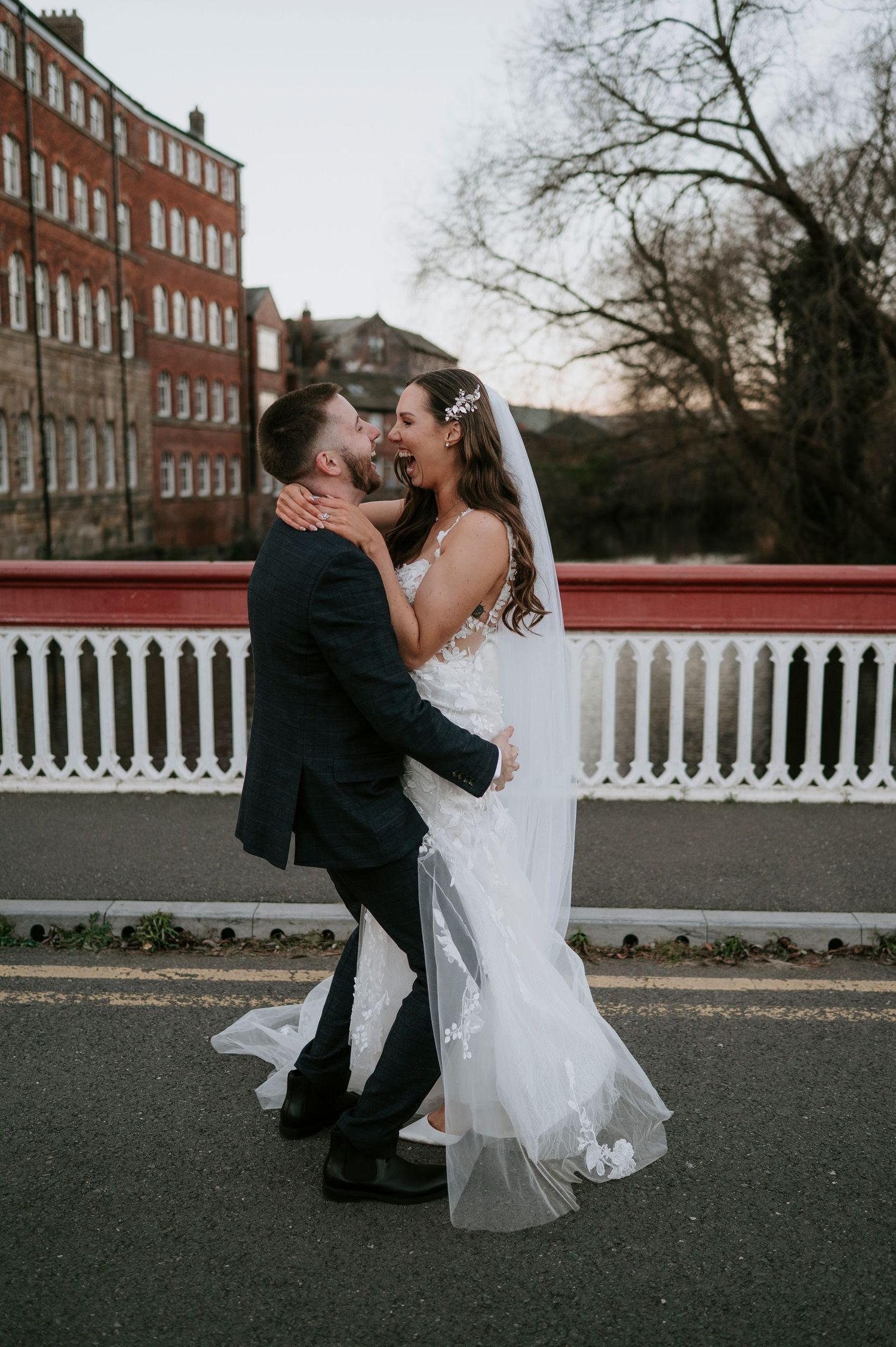 Bride and groom share a romantic embrace on a bridge, with red brick buildings in the background.