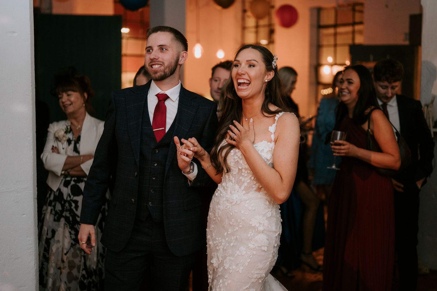 Smiling bride and groom entering wedding reception to cheers from gathered guests.