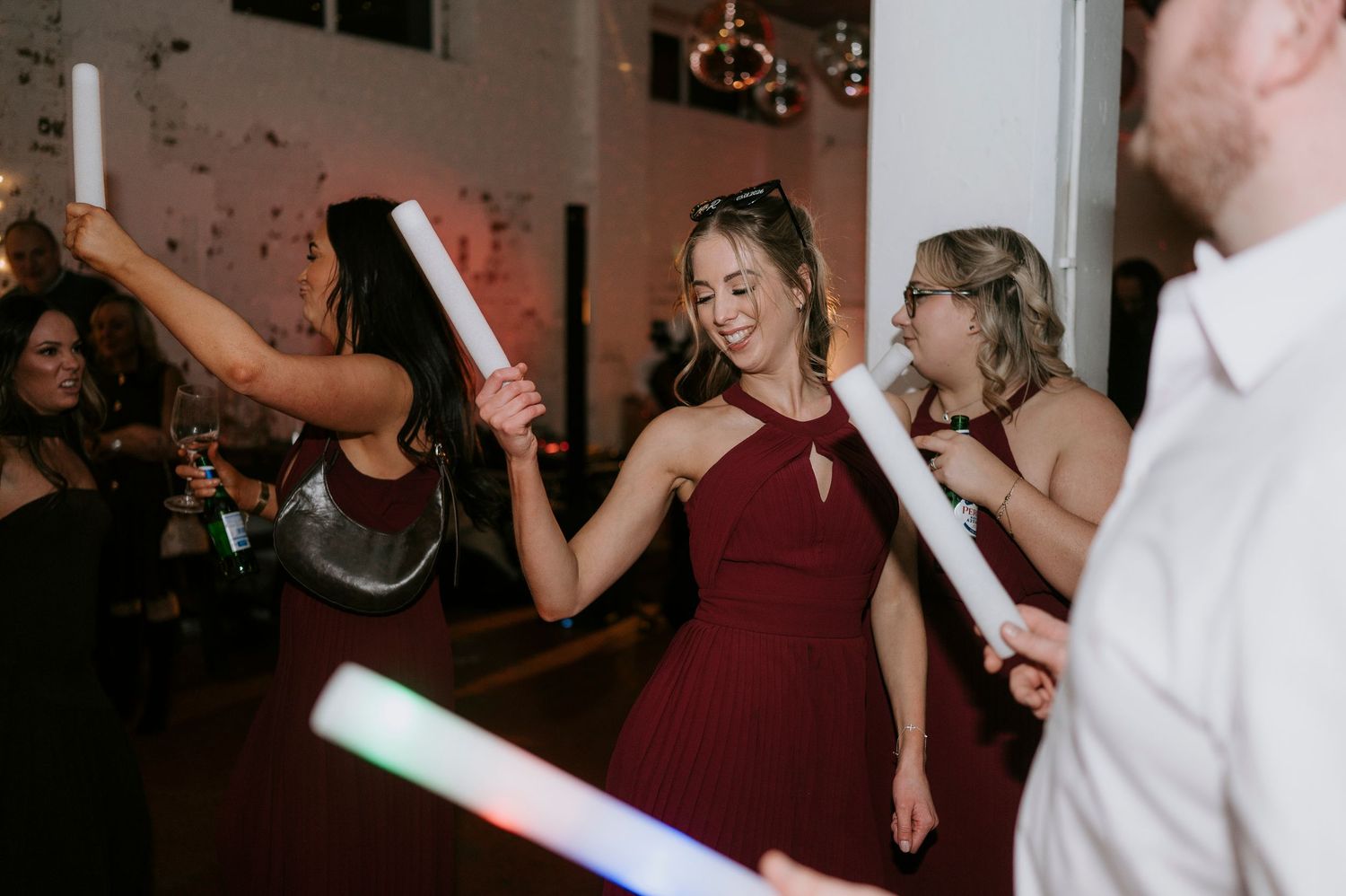 Bridesmaids in burgundy dresses dancing with glow sticks at a wedding reception.