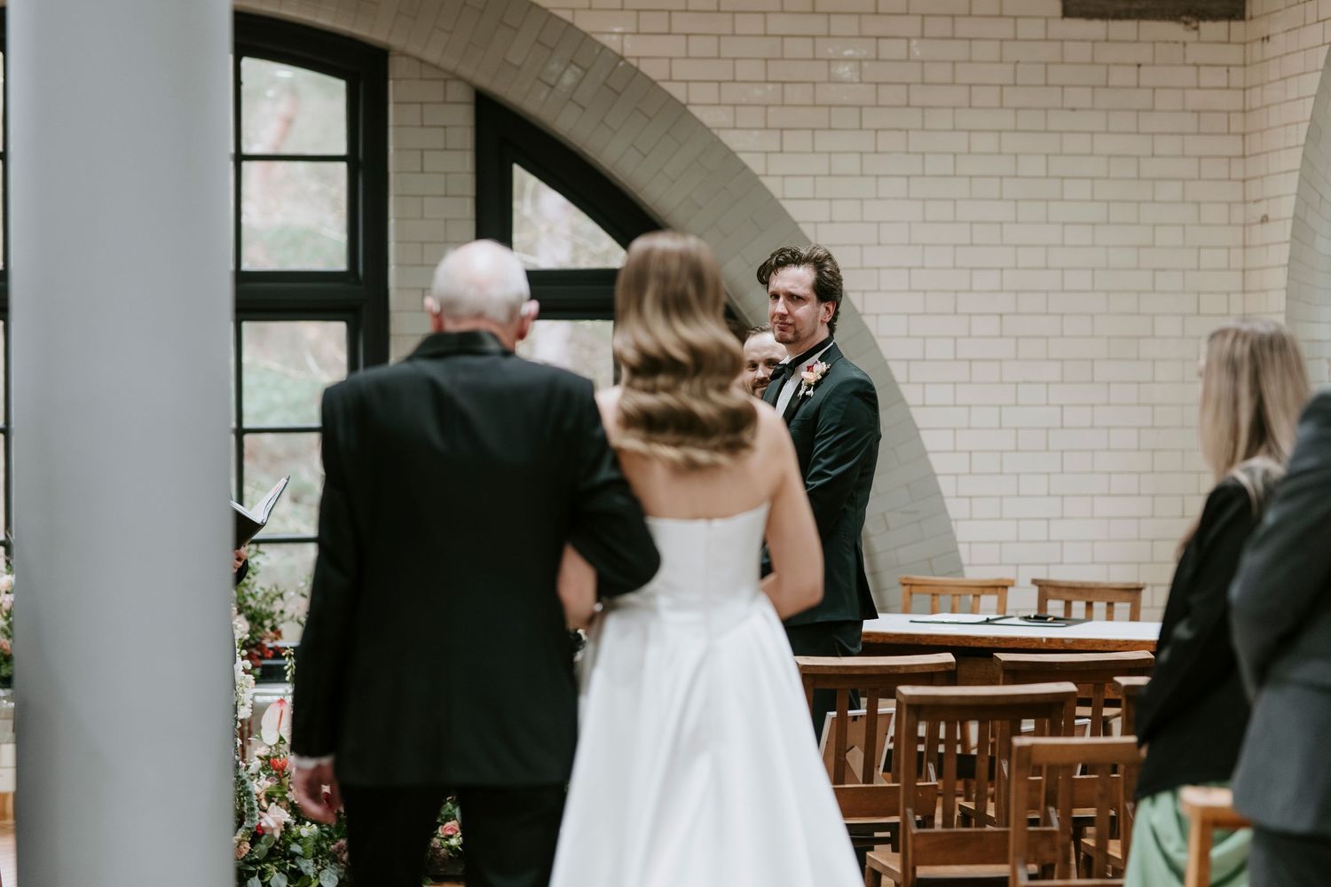 A wedding ceremony takes place in an indoor venue with white brick walls and wooden chairs.