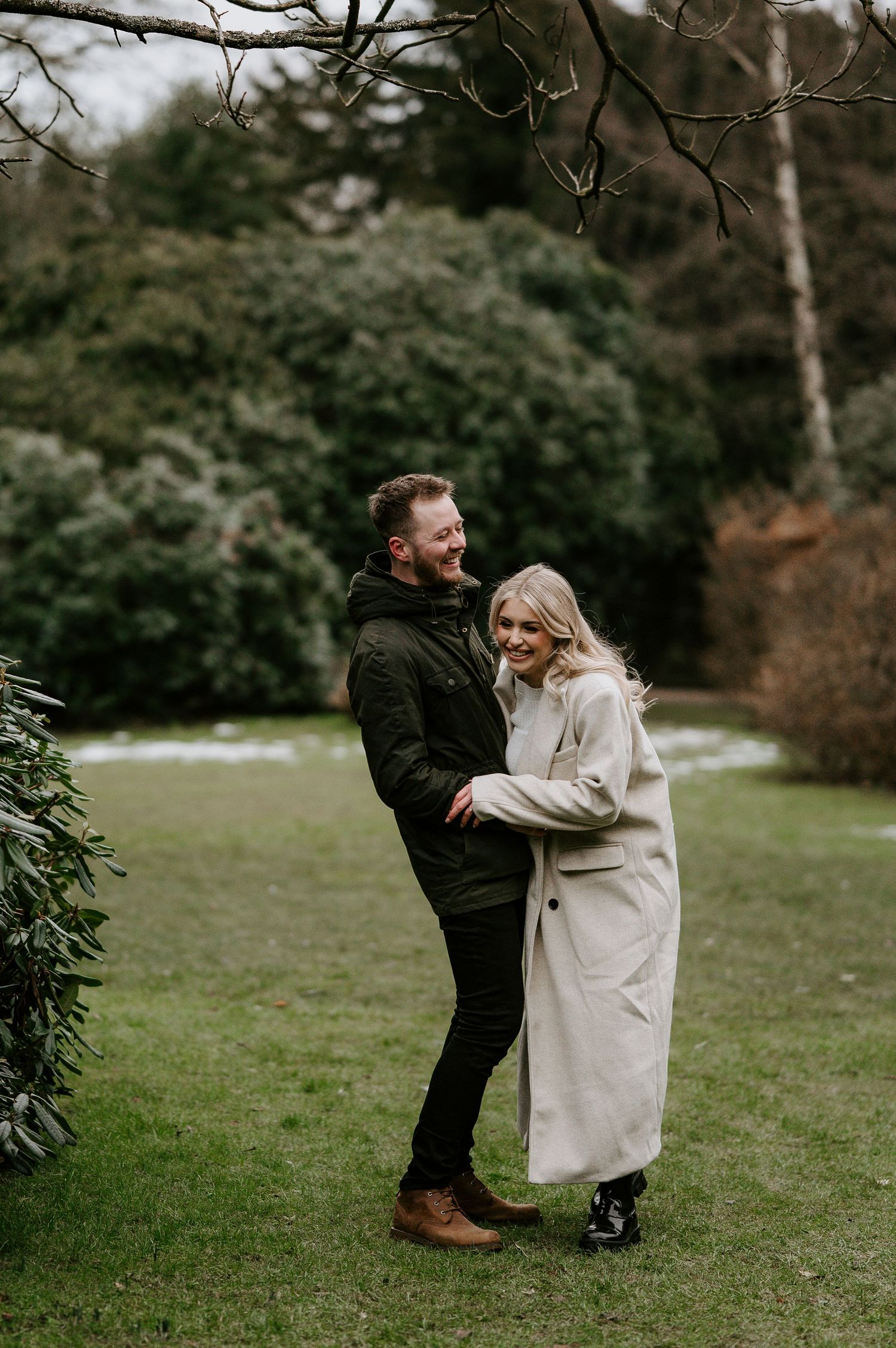 A couple in winter attire shares playful moments in a park setting during a romantic outdoor photoshoot.