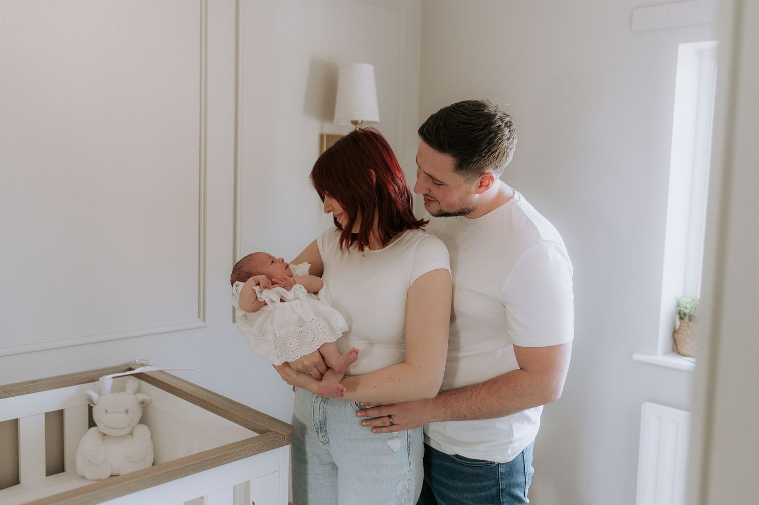 Parents stand together holding their newborn baby in a bright nursery room next to a white crib.