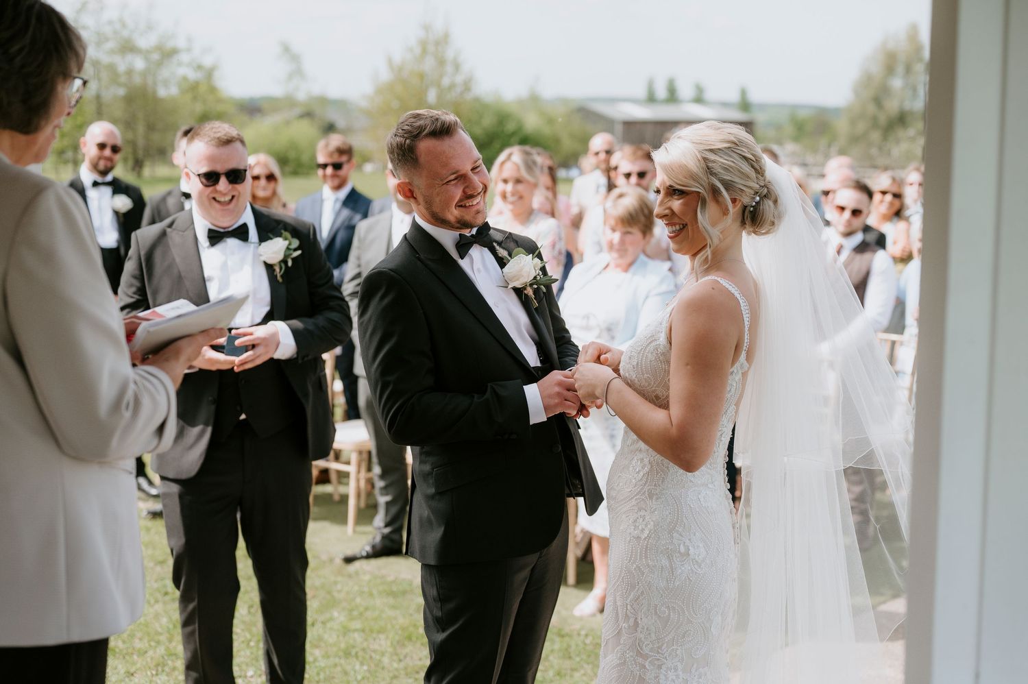 A couple exchanges rings during an outdoor wedding ceremony surrounded by guests on a sunny day.