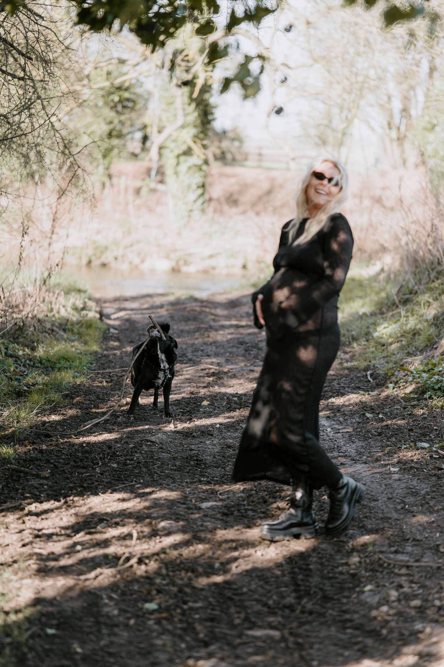 A figure in dark clothing walks with a black dog along a shaded woodland path on a sunny afternoon.