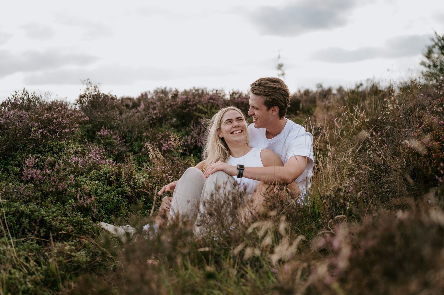 A romantic couple embraces in a golden field during sunset, surrounded by tall grass and wildflowers.