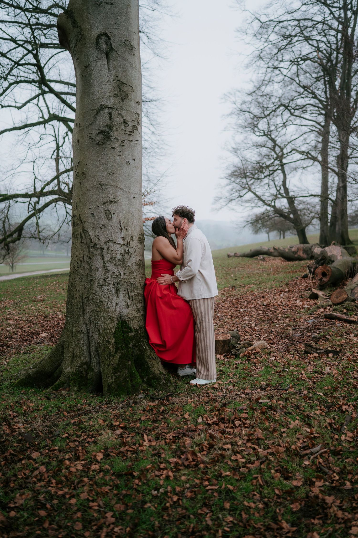 A couple shares tender moments among tall beech trees in a misty autumn forest with fallen leaves on the ground.