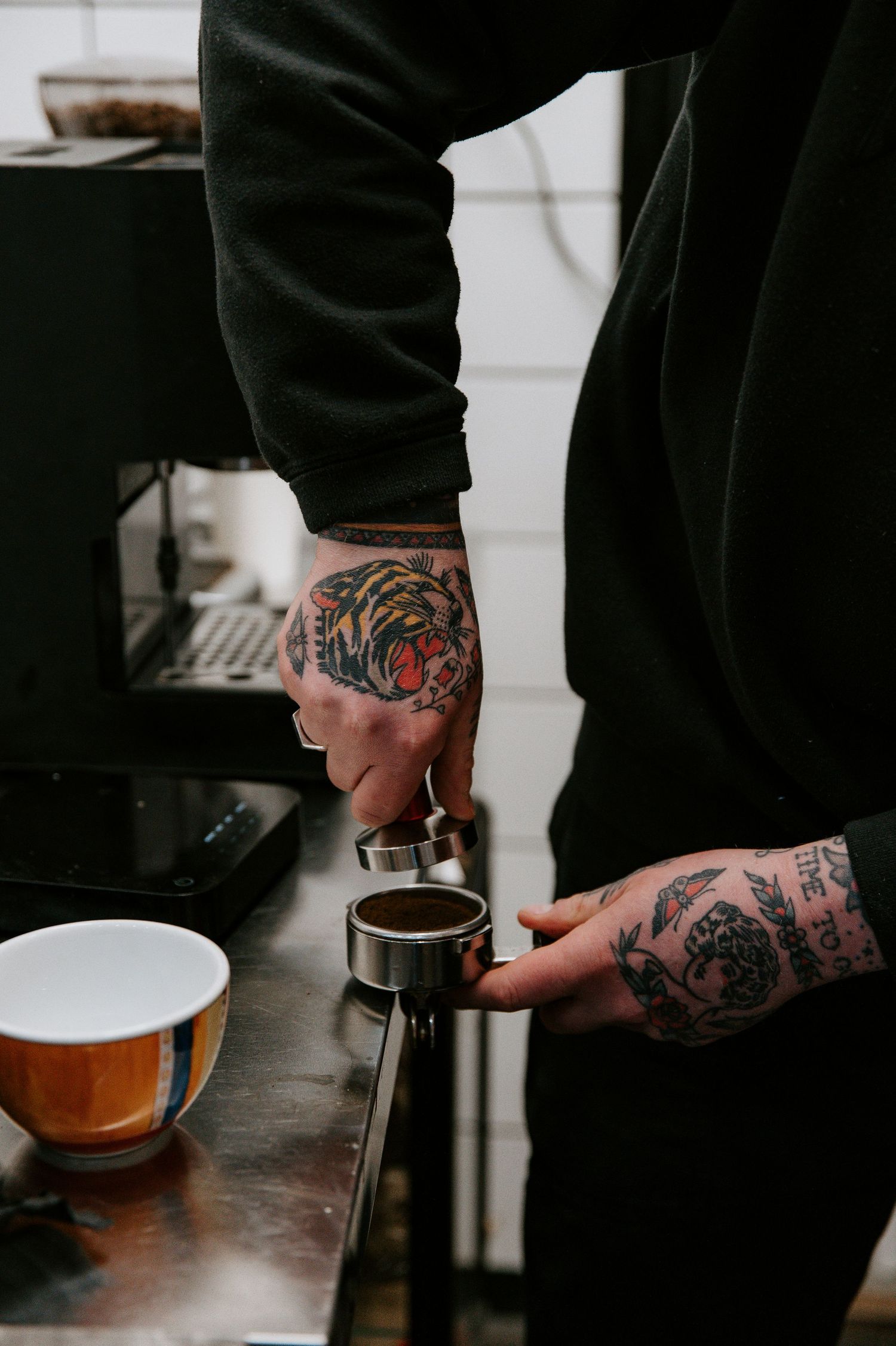 Tattooed hands prepare coffee using professional equipment in a dark setting with multiple brewing steps shown.