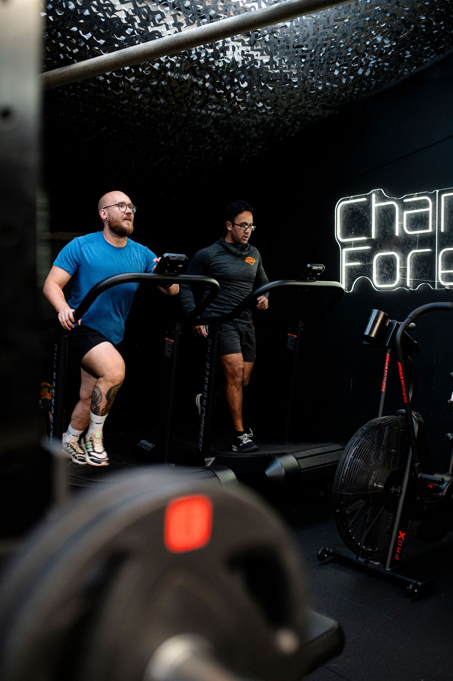 Two people working out on exercise equipment in a dark gym with illuminated 'Change Force' sign on wall.