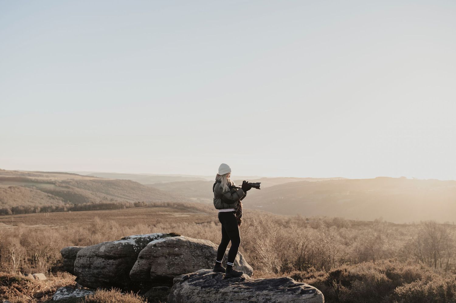 A photographer stands on a rocky outcrop with camera gear during golden hour in a hilly landscape.