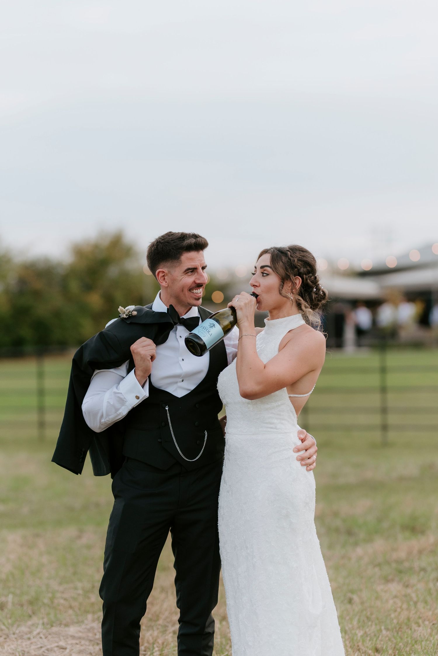 A playful wedding couple takes turns drinking from champagne bottles in an outdoor field setting.