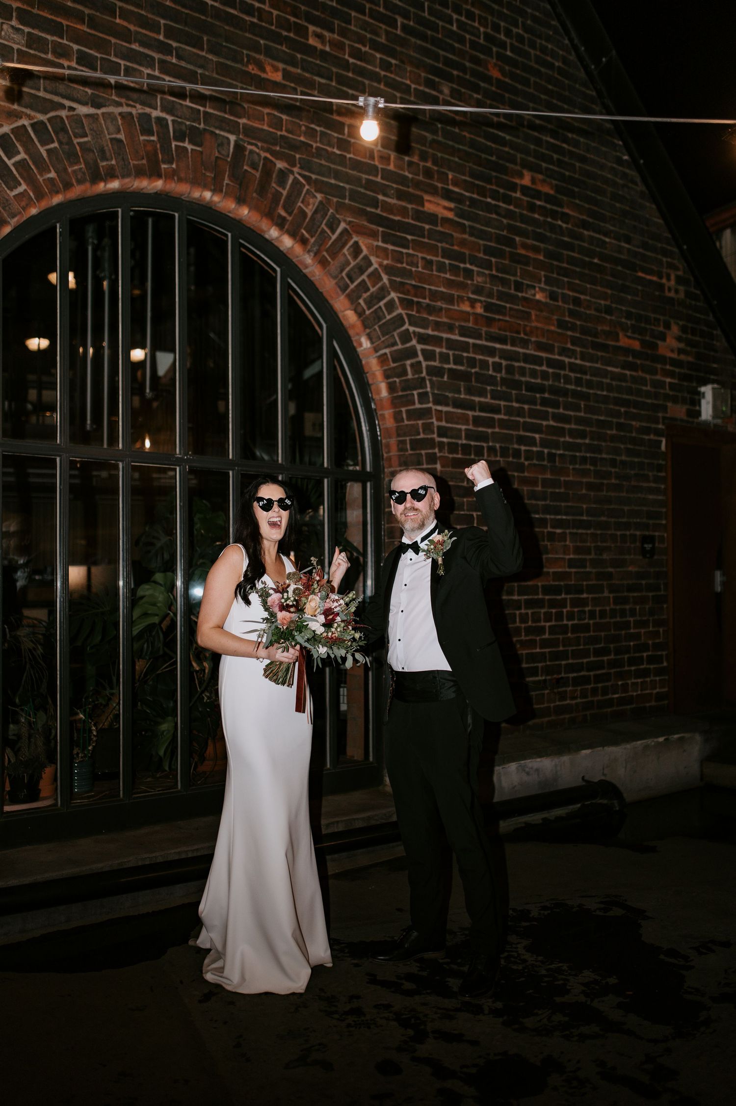 Wedding couple poses playfully with sunglasses against brick wall at night.