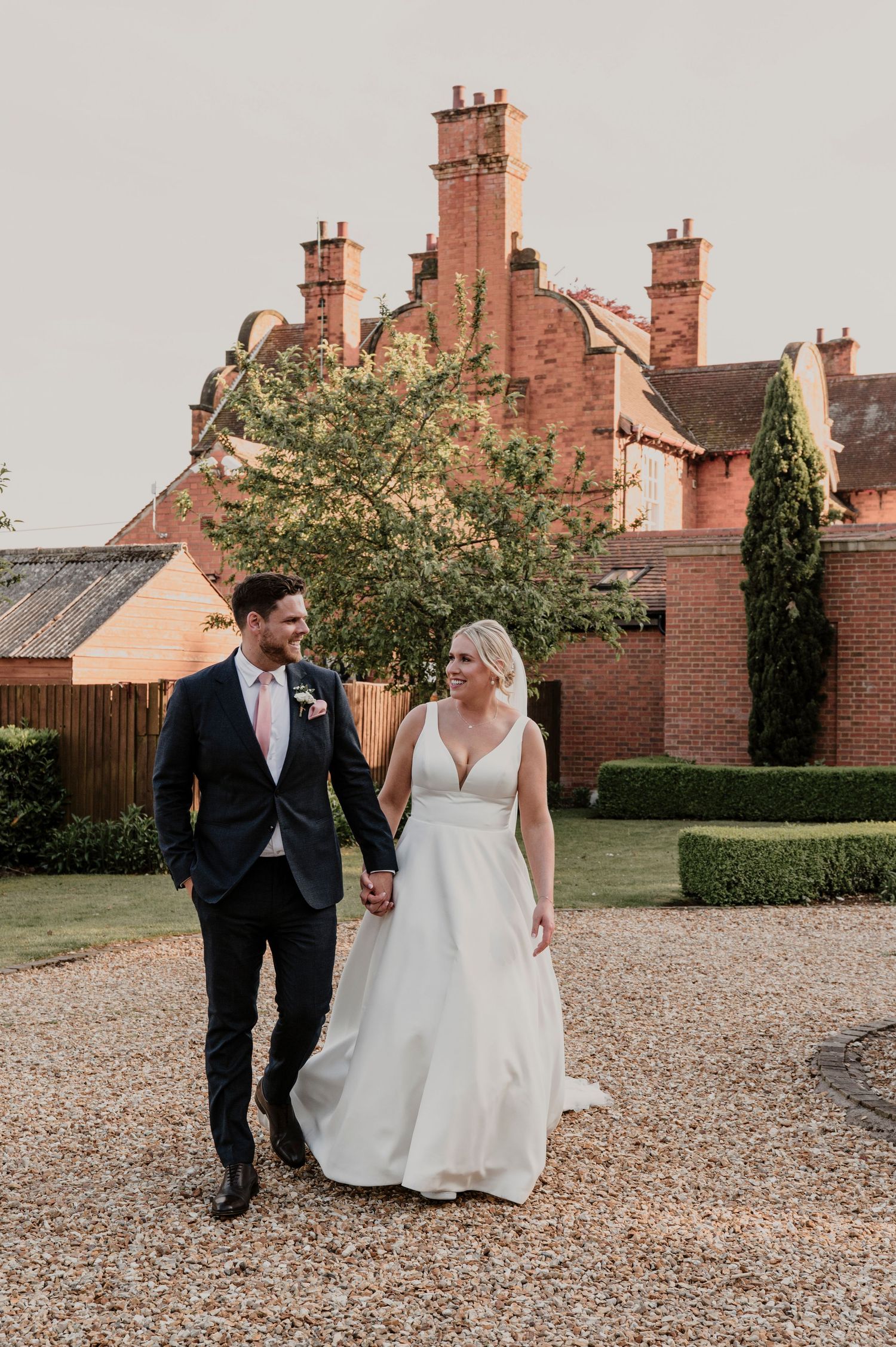 Couple walking together on gravel path with red brick buildings and greenery in background.