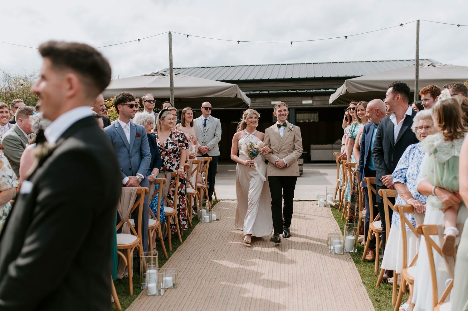 Wedding guests stand in rows outdoors during a summer ceremony with wooden chairs and floral decorations.
