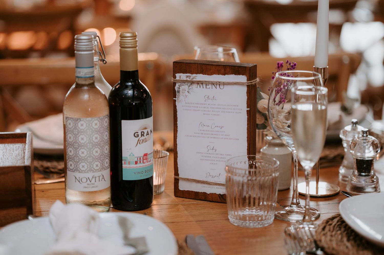 Elegant table settings with wine bottles and glassware arranged for a rustic wedding reception in warm lighting.