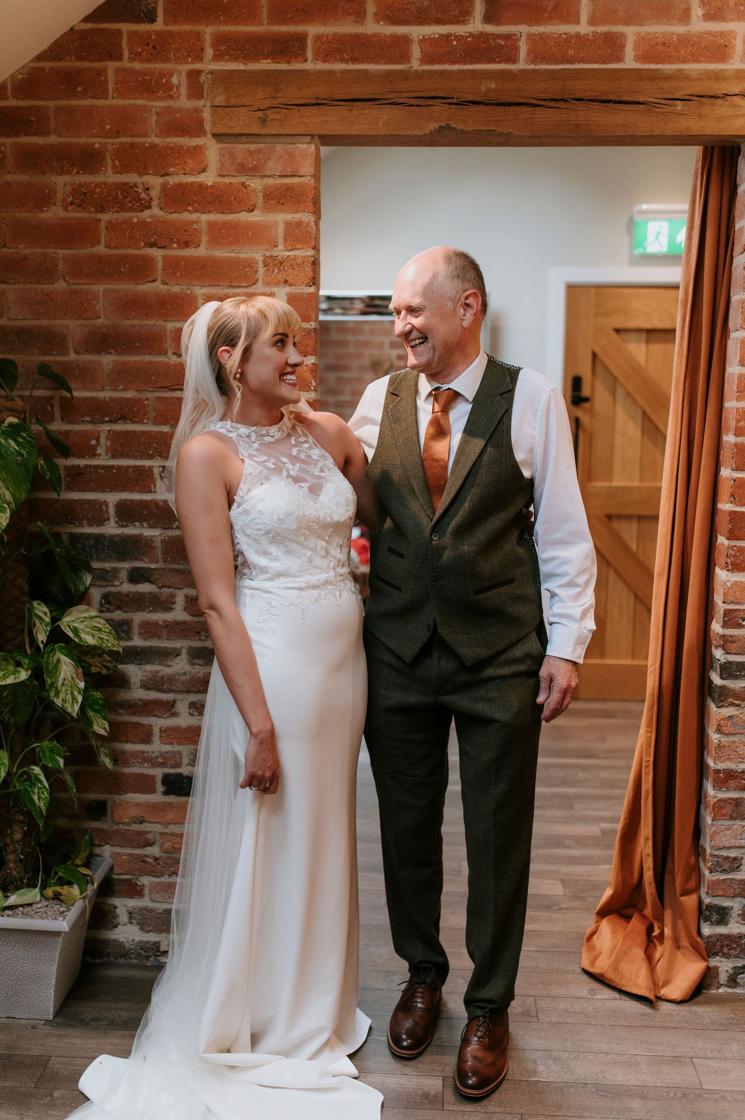 Couple in wedding attire share a joyful moment against exposed brick wall in indoor venue.